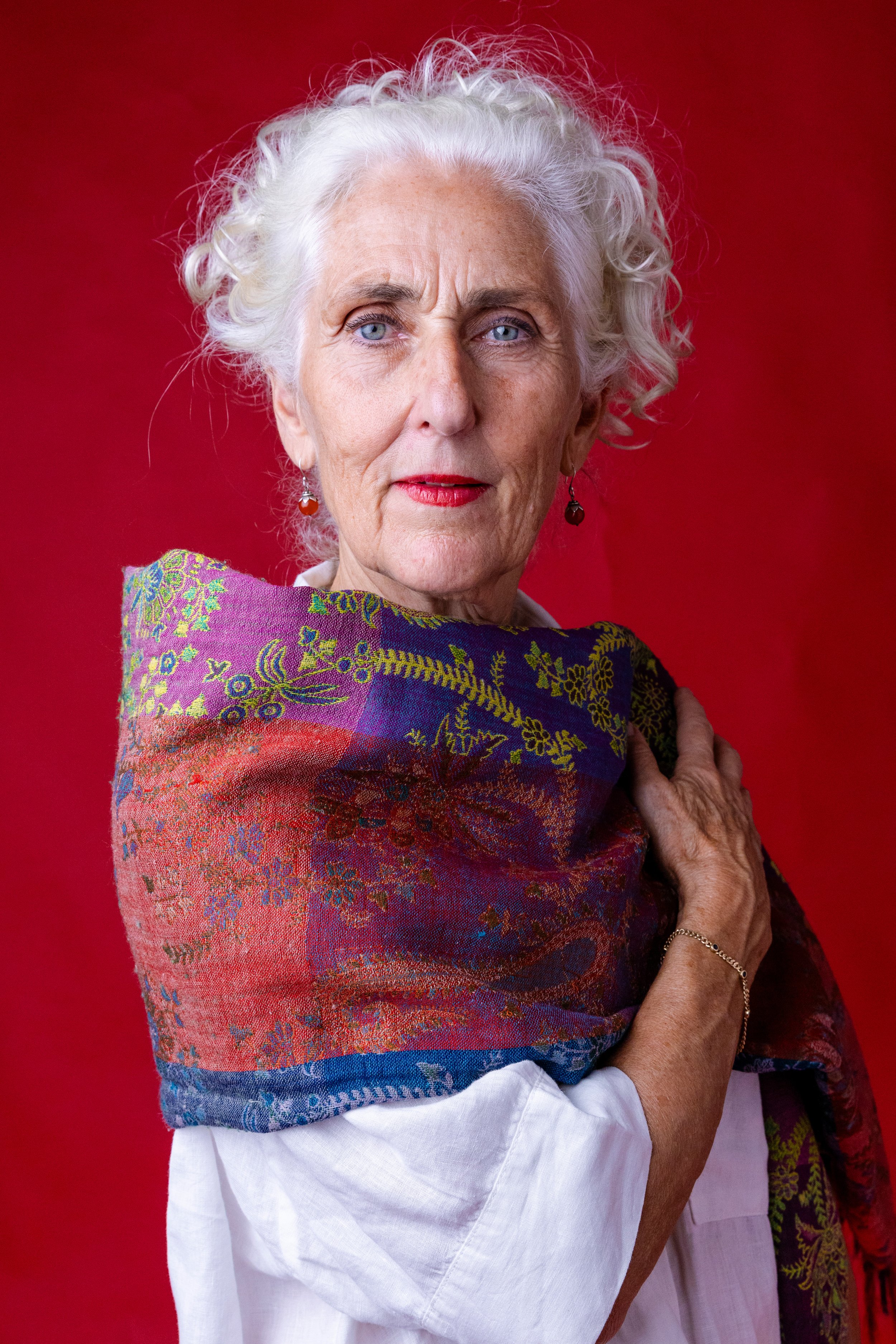 An elderly woman with gray, curly hair and blue eyes wearing a colorful shawl and a white blouse against a red background.