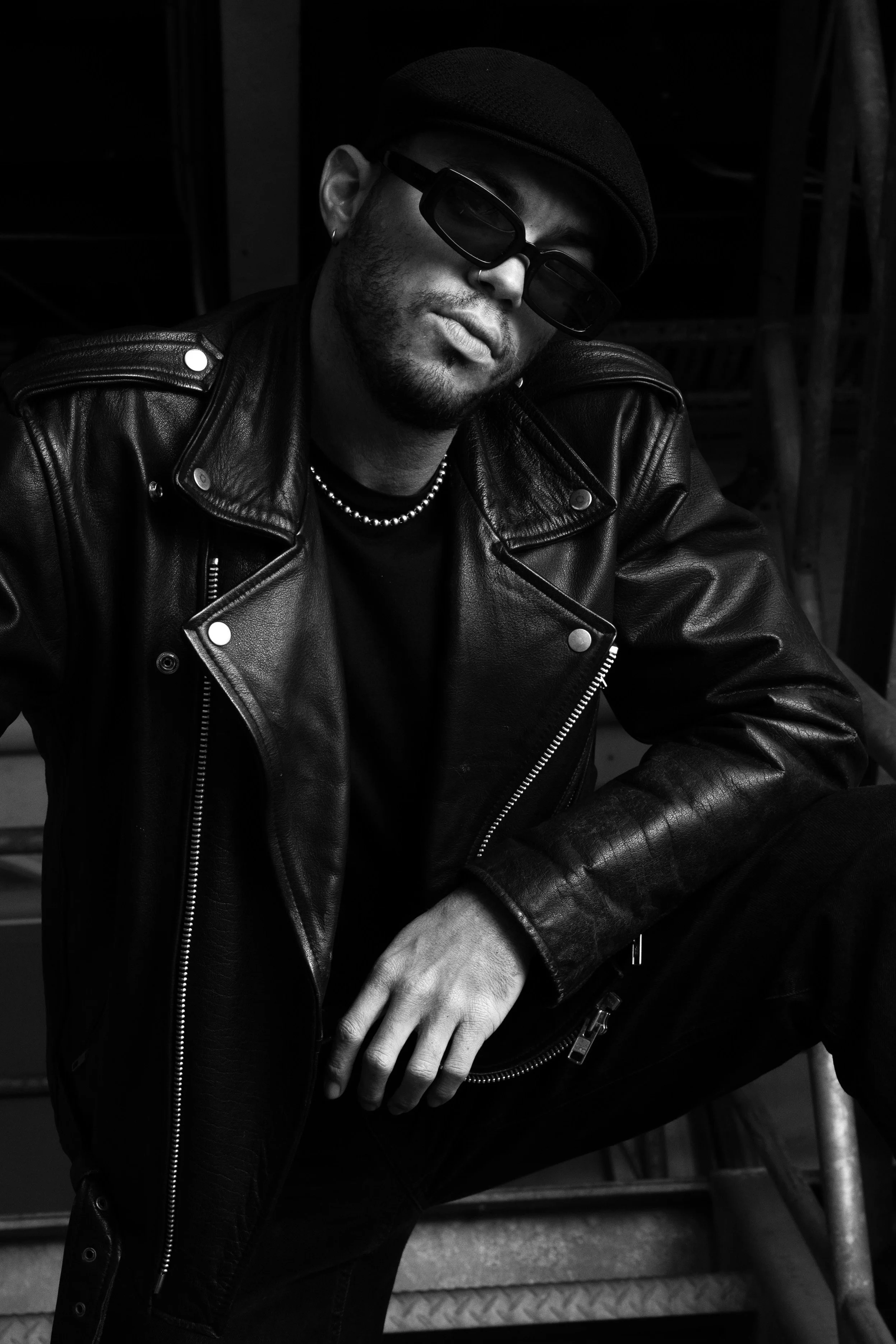 A man wearing sunglasses, a beanie, a leather jacket, and a beaded necklace, sitting in a dark setting with a serious expression.