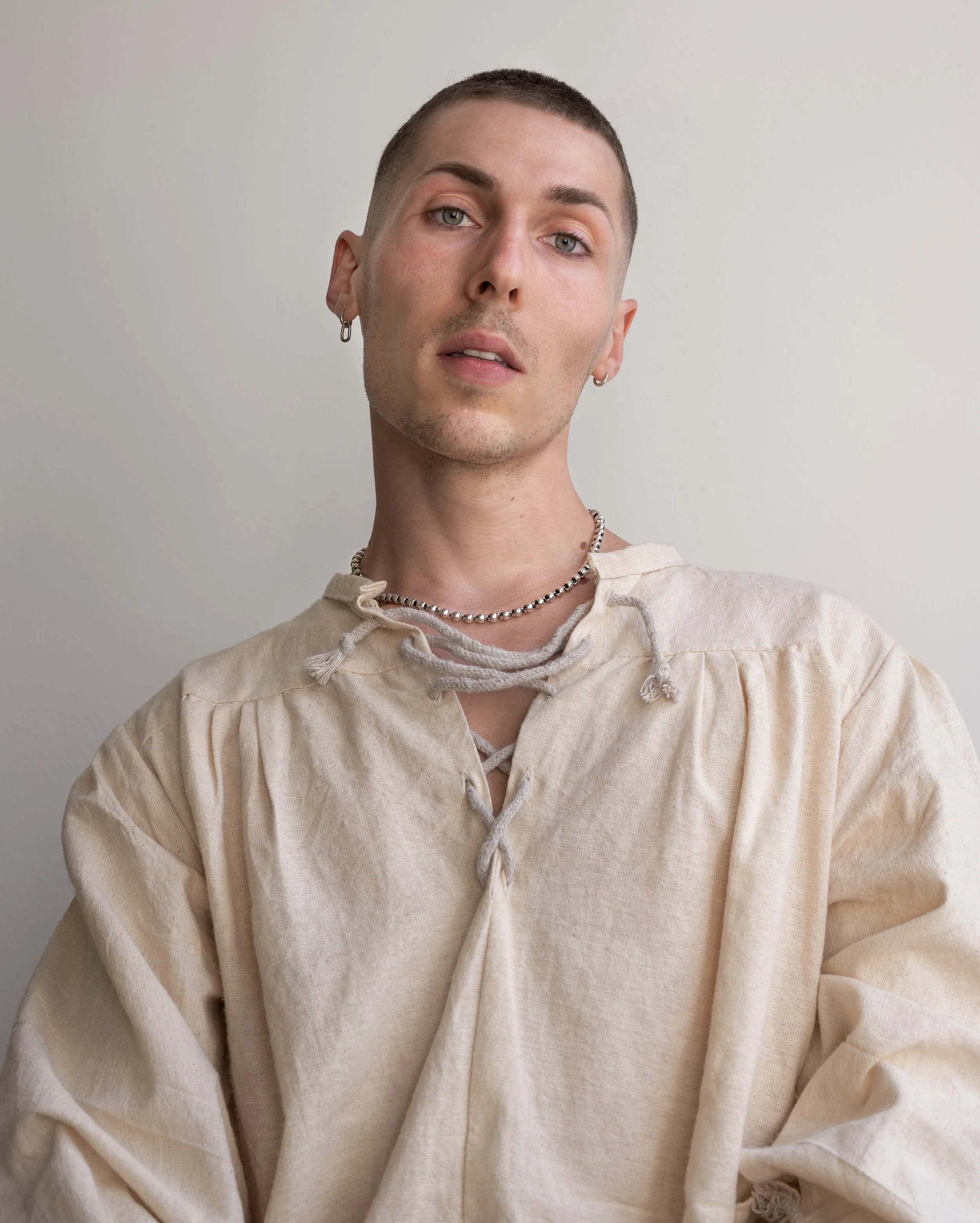A young person with short hair, earrings, a necklace, wearing a beige, slightly distressed shirt with laces at the collar, posing against a plain light background.