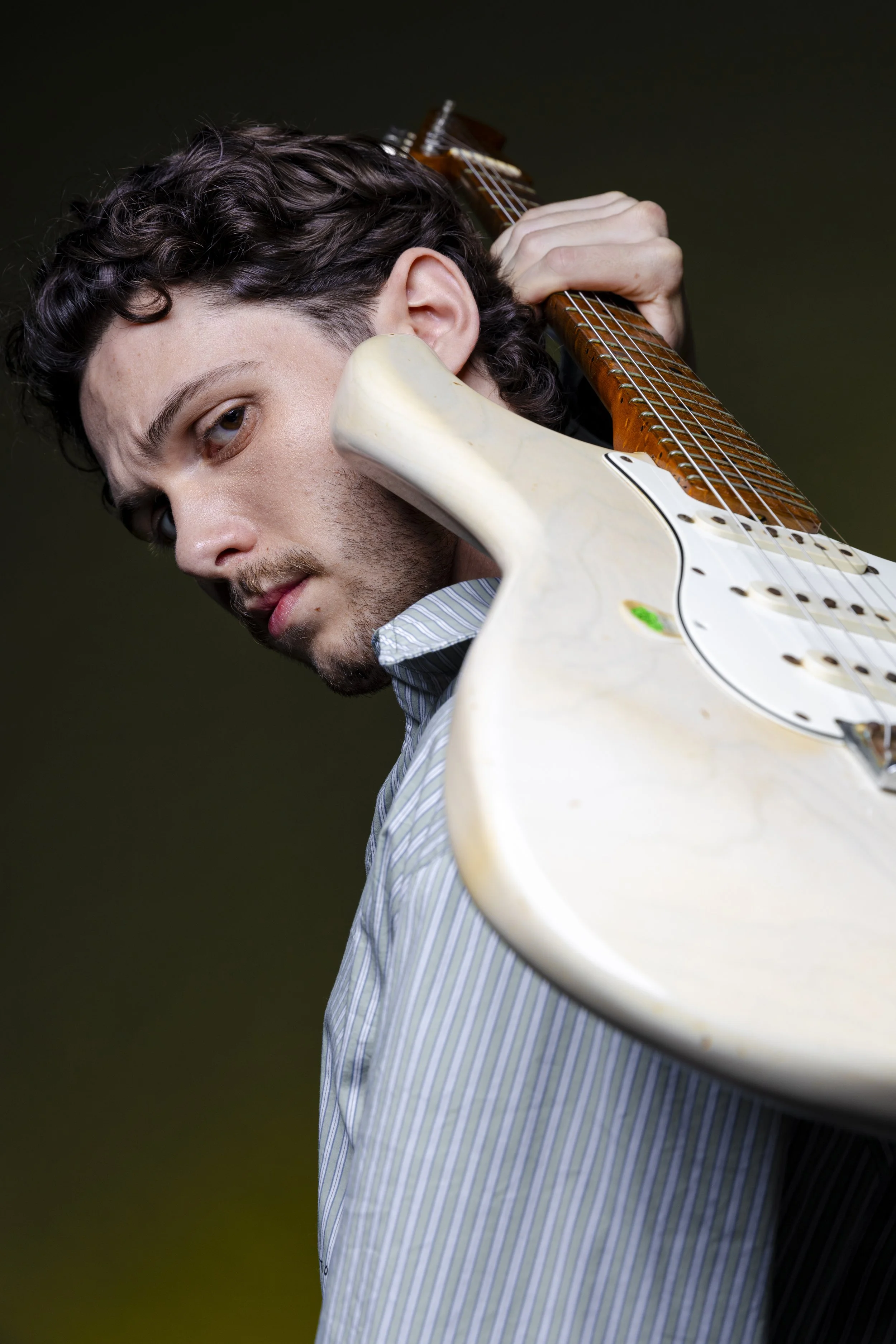 A young man with curly dark hair and light skin holding an electric guitar over his shoulder, looking at the camera with a confident expression.
