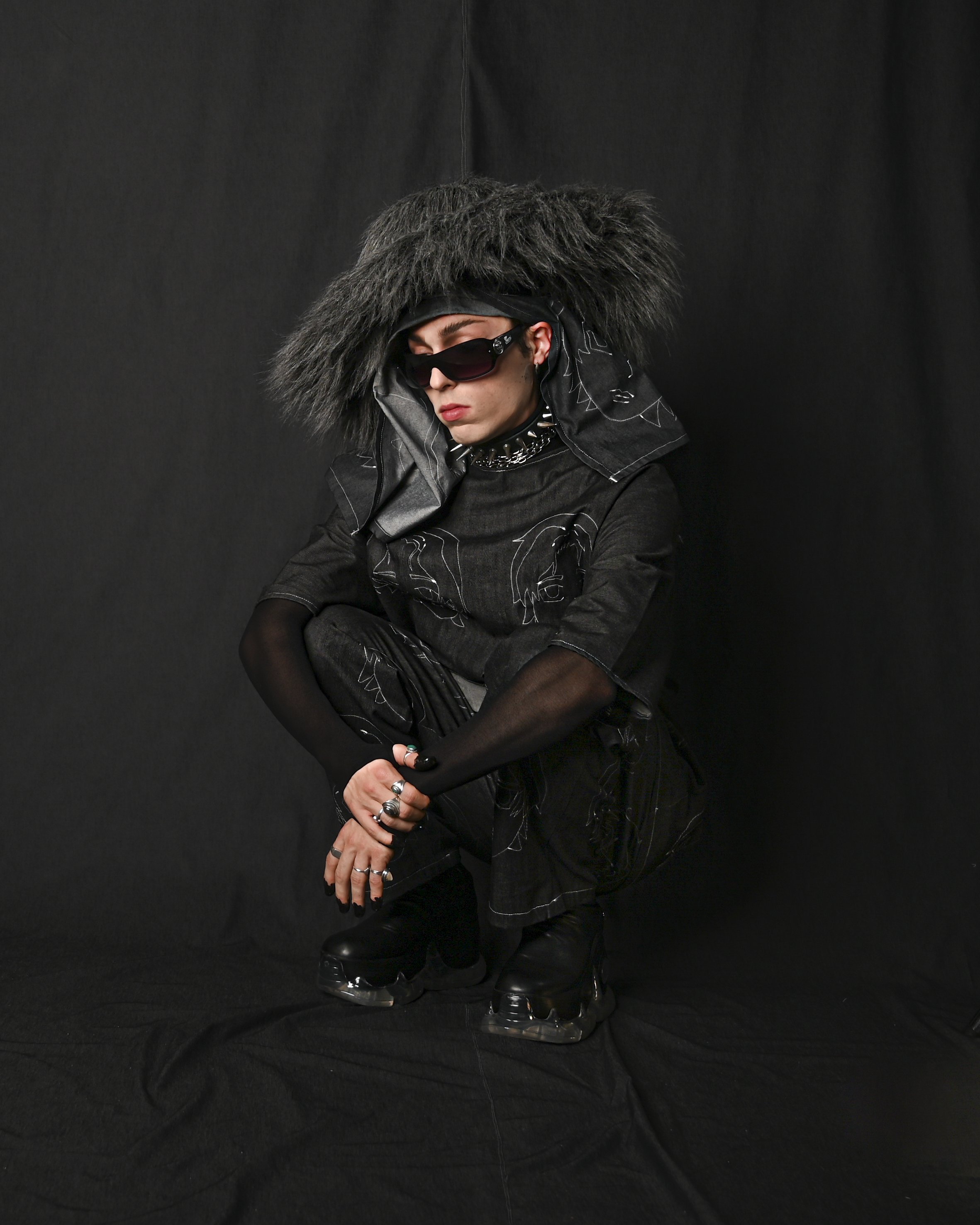 Person dressed in all black with oversized black furry hat, sunglasses, multiple rings, and platform shoes, crouching against a black background.
