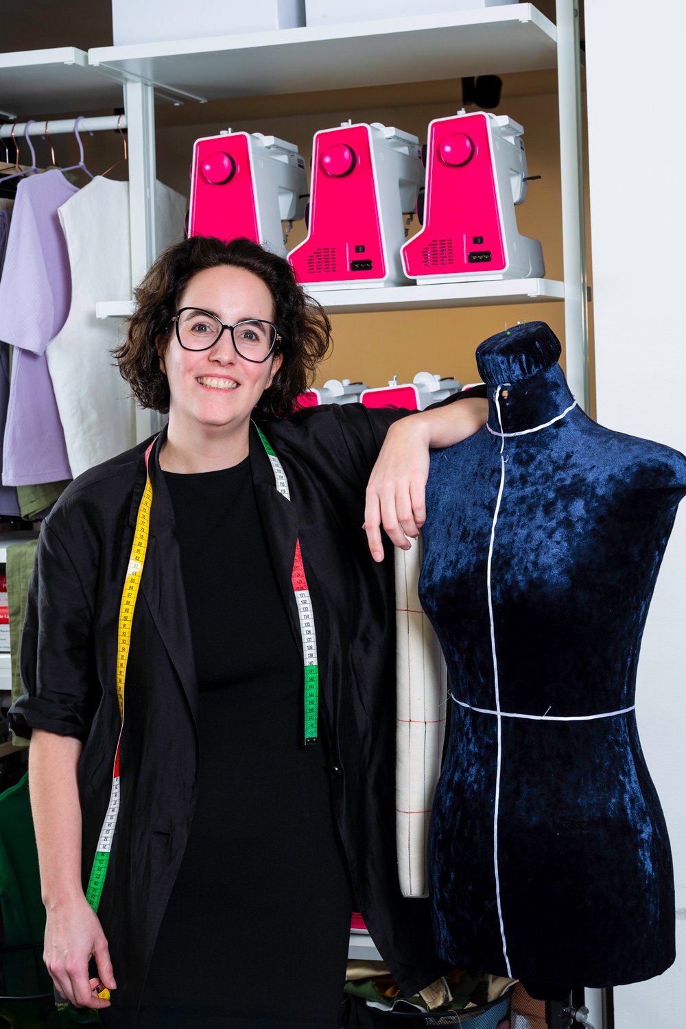 A woman with glasses smiling, standing next to a dress form covered with dark fabric, in a workspace. She has a measuring tape draped around her neck and is surrounded by sewing equipment and fabric.