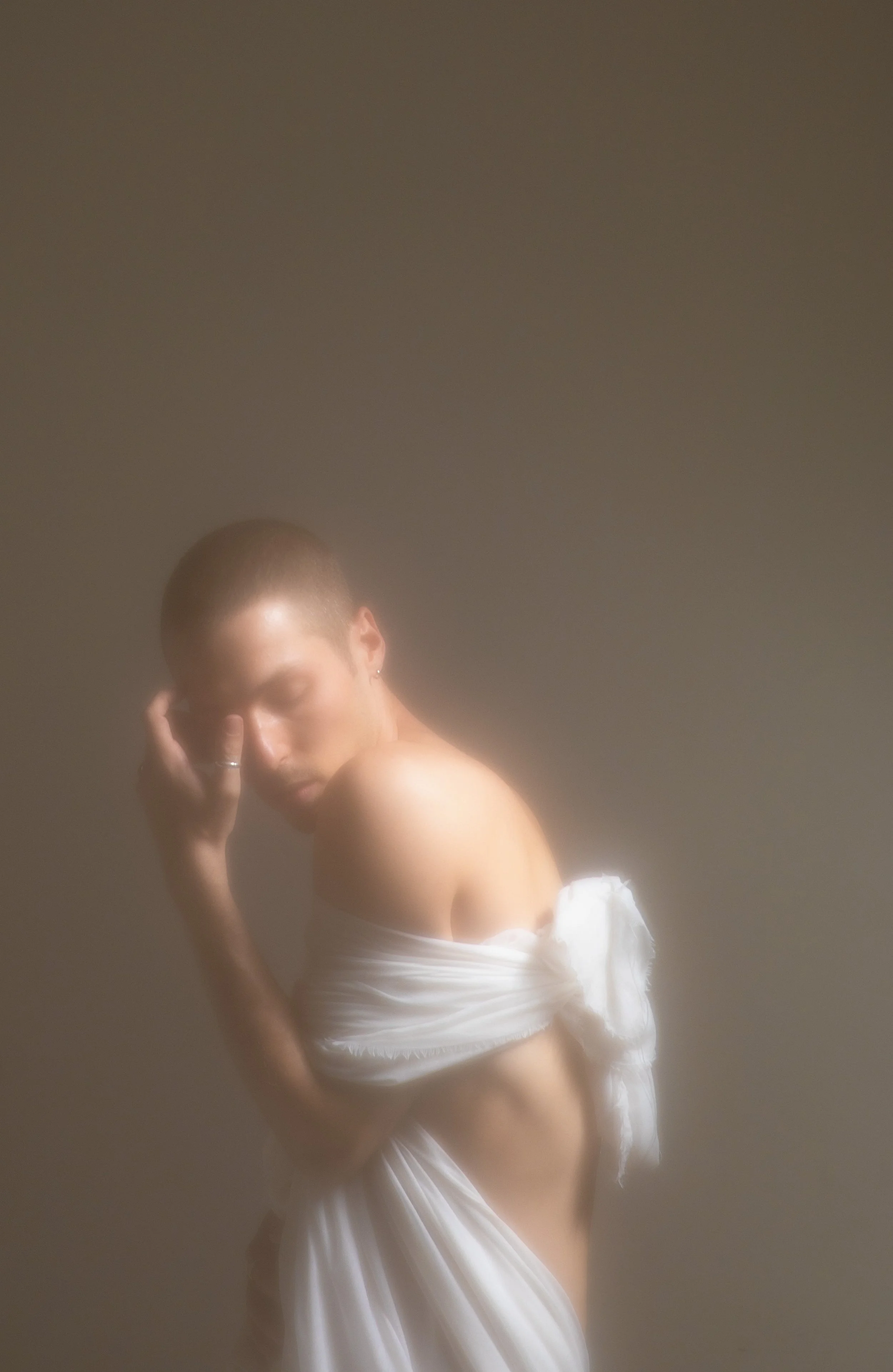 A woman with a shaved head wrapped in white fabric, posing with her eyes closed, against a soft, blurred background.