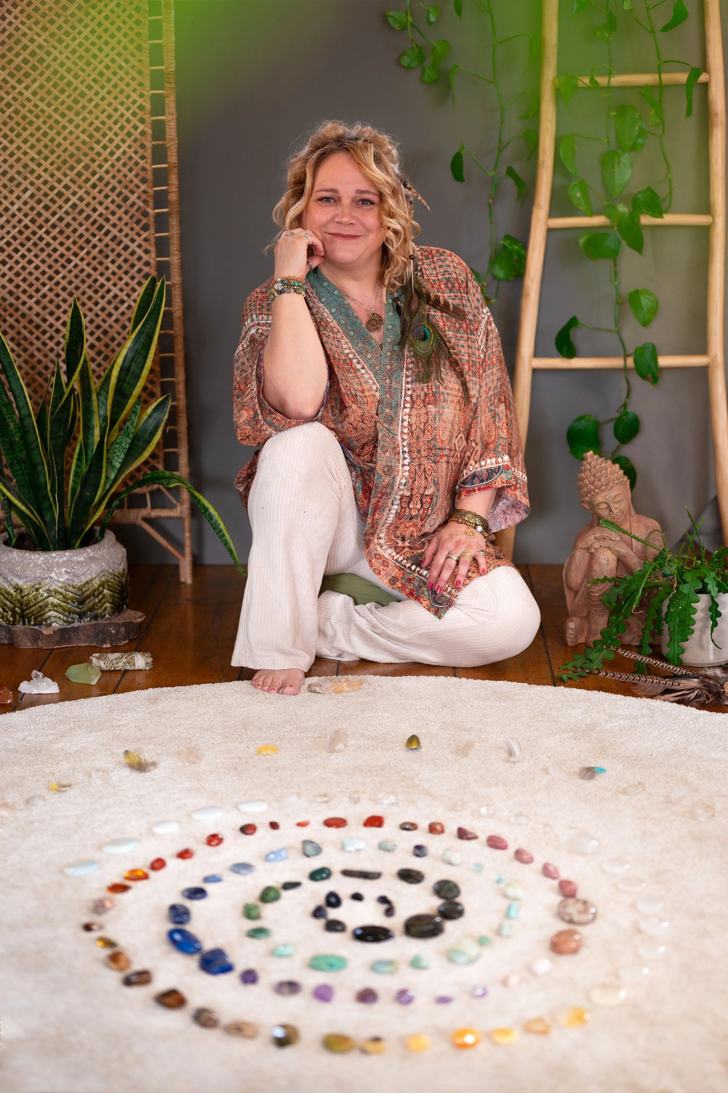 A woman with curly blonde hair sits cross-legged on a carpet, smiling. She wears a patterned blouse and white pants. Behind her are green plants, a wooden ladder, and a Buddha statue, with a spiral arrangement of various colored stones on the carpet 