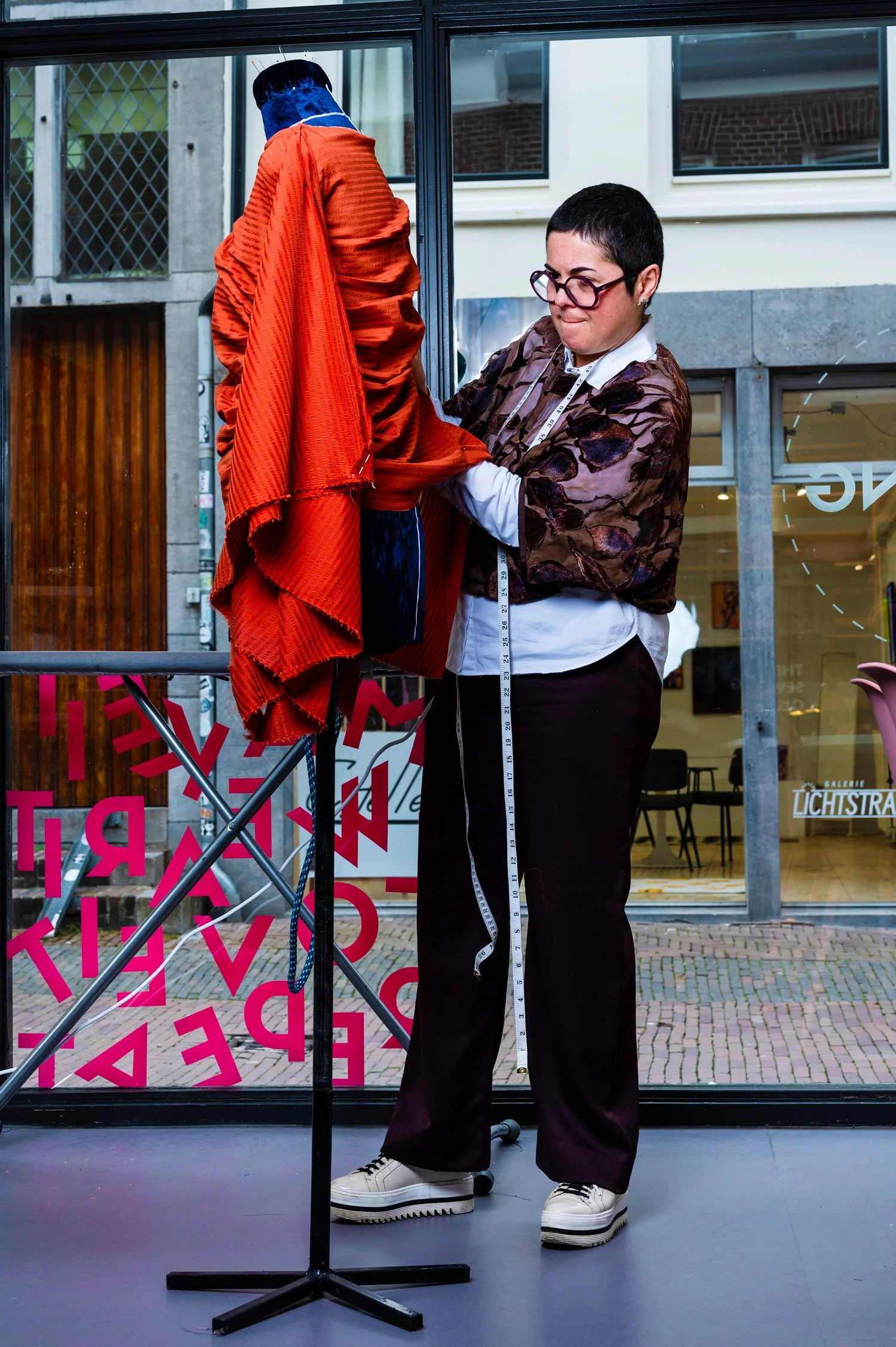 A woman with short dark hair and glasses measuring a red fabric on a dress form outside a modern building with large glass windows.