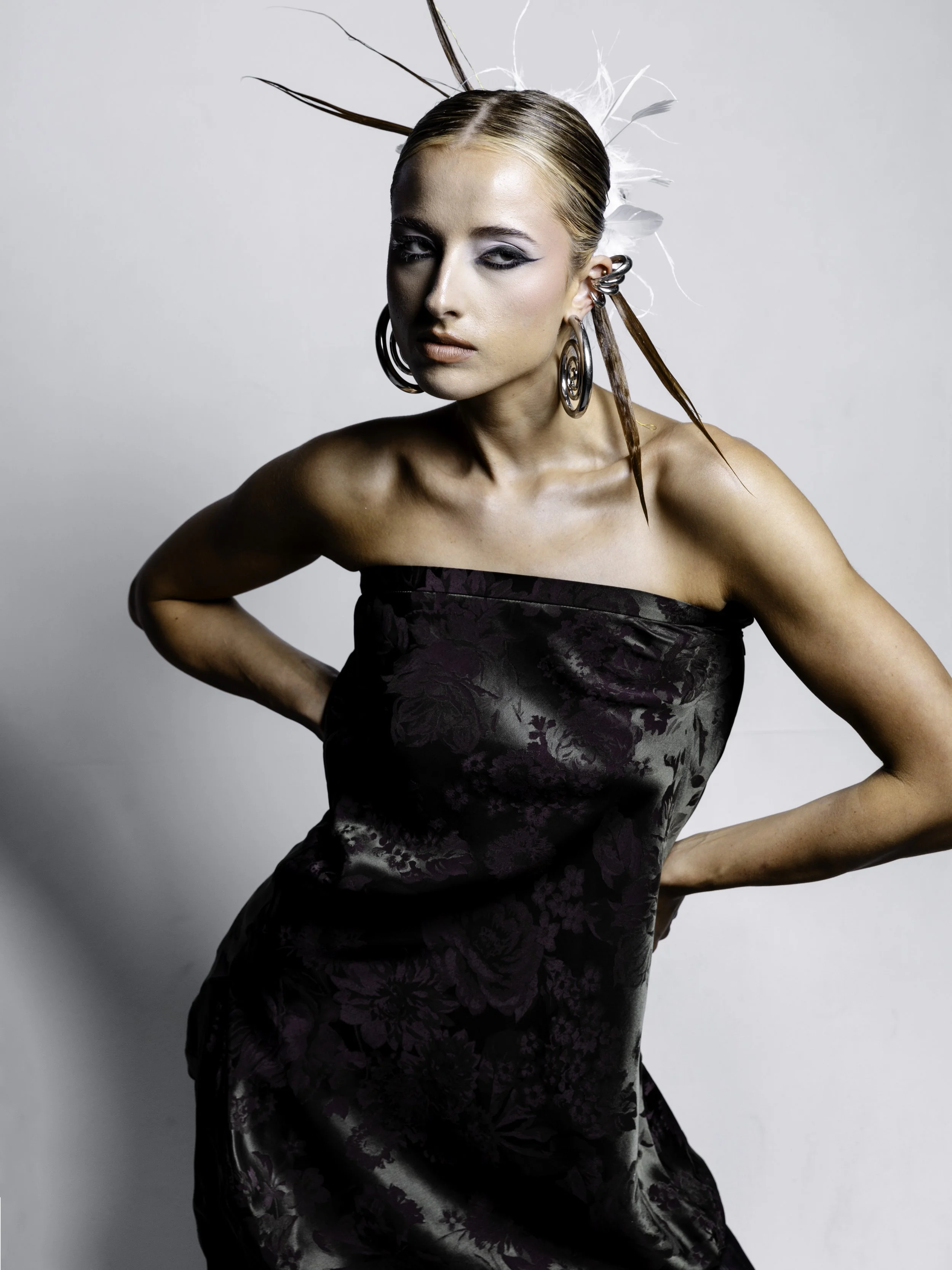 A woman with styled hair, dramatic eye makeup, and large hoop earrings posing in a strapless black floral dress against a plain light background.
