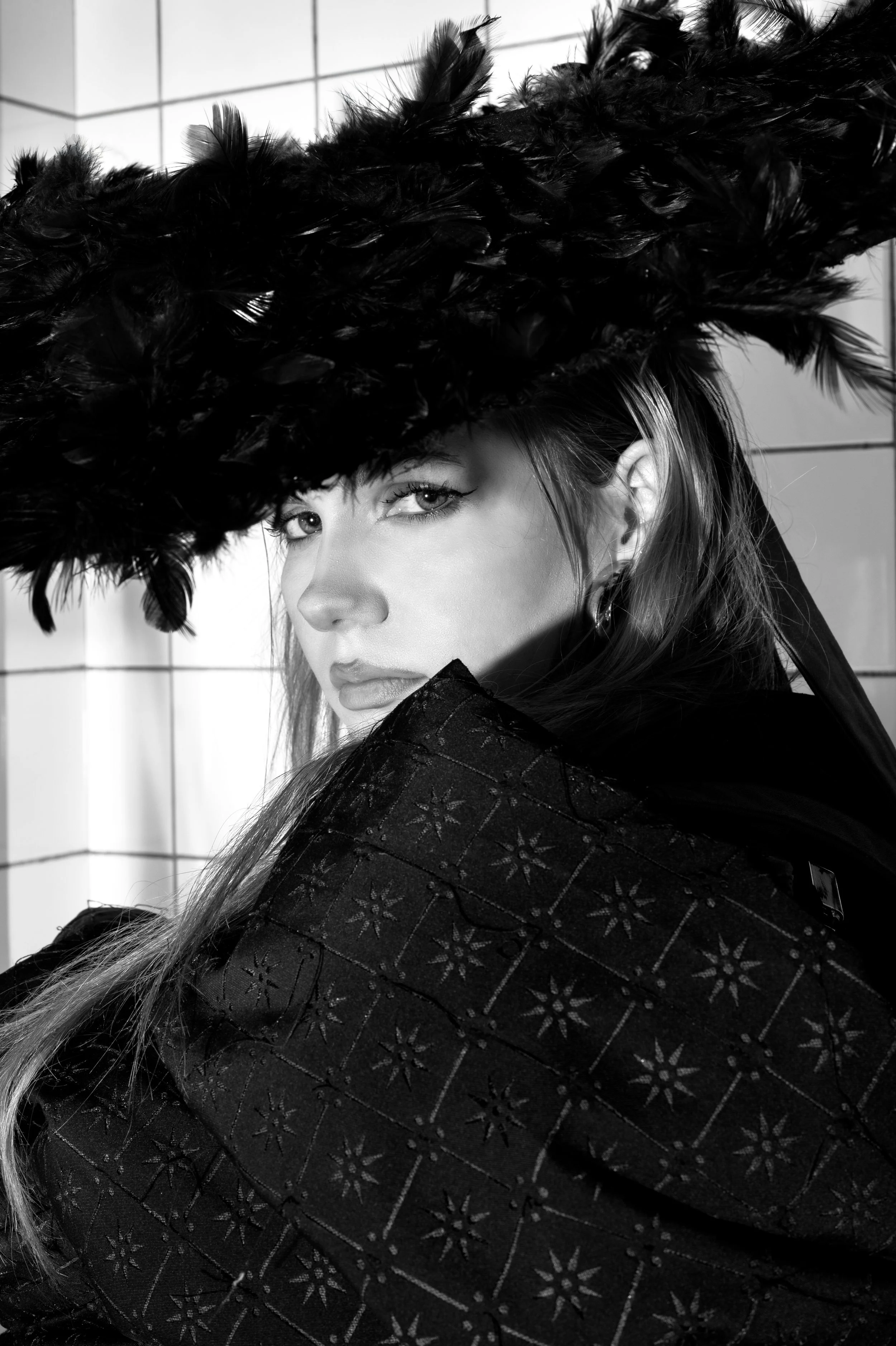 A woman wearing a large feathered hat and a patterned coat, looking over her shoulder in a black and white photograph.