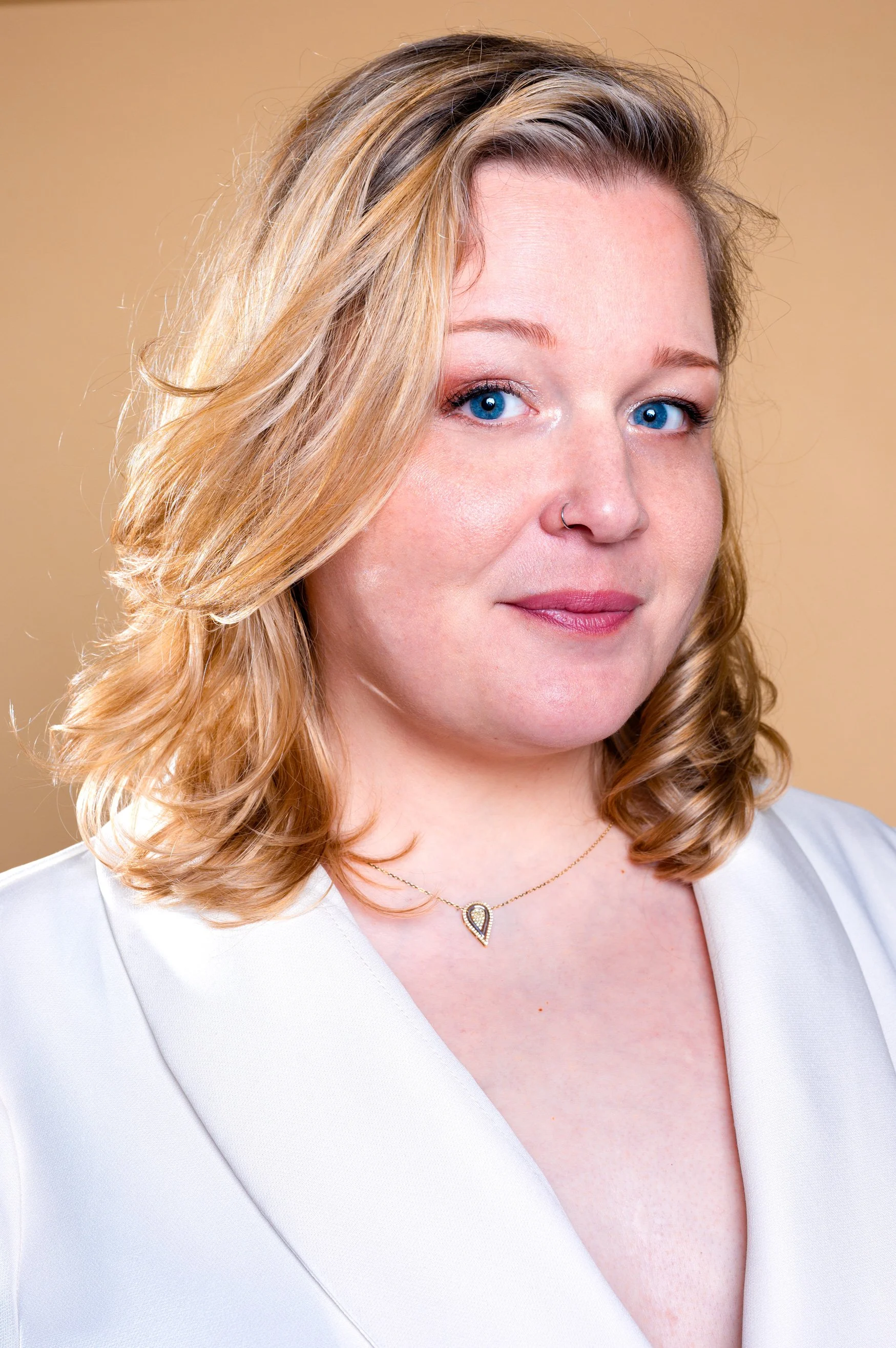 A woman with blonde, shoulder-length hair, blue eyes, and a nose piercing, wearing a white blazer and a gold necklace with a pendant, standing against a beige background.