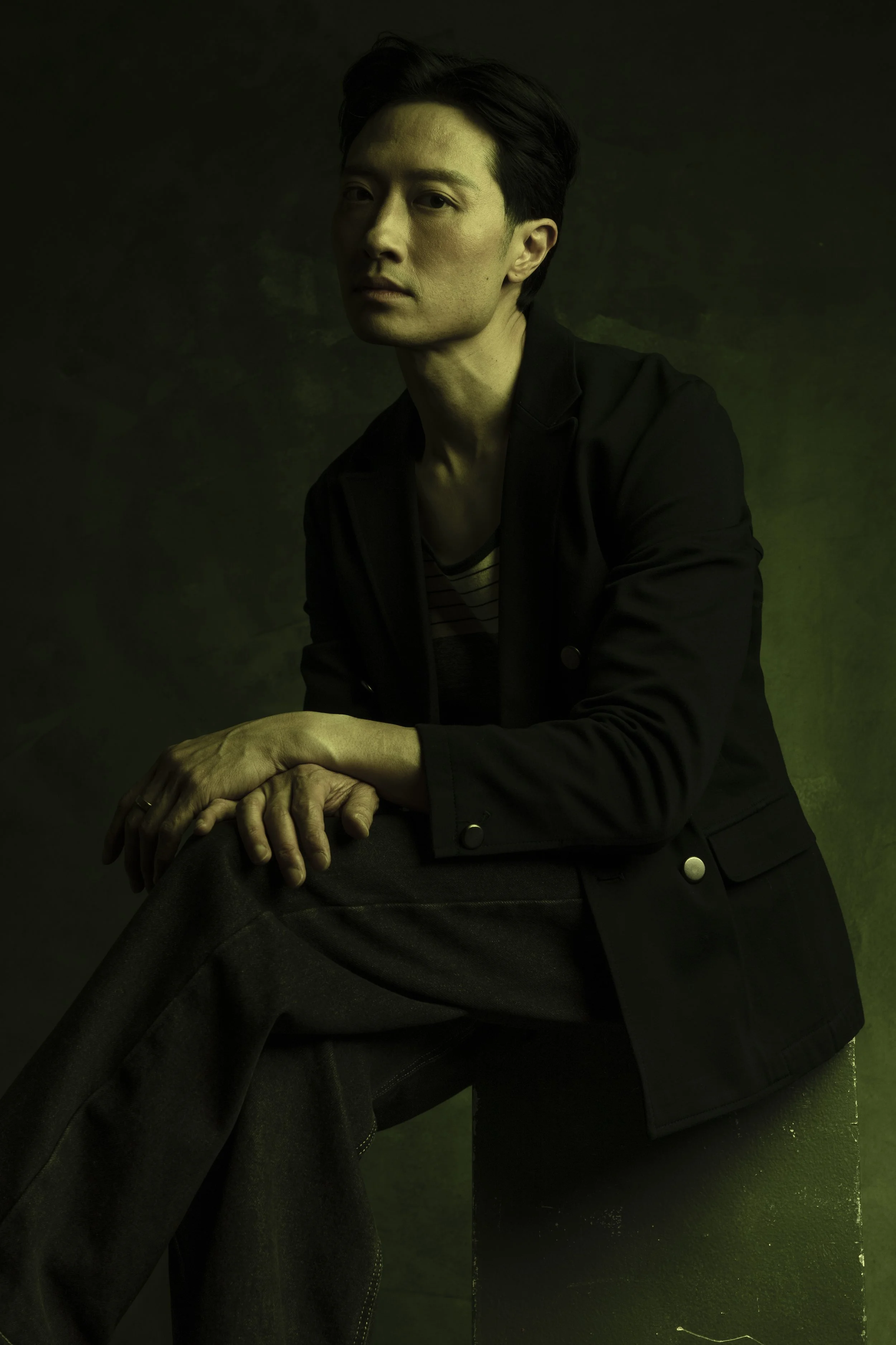 A man with dark hair, wearing a black jacket, striped shirt, and dark pants, seated on a black stool against a dark green background, looking at the camera.