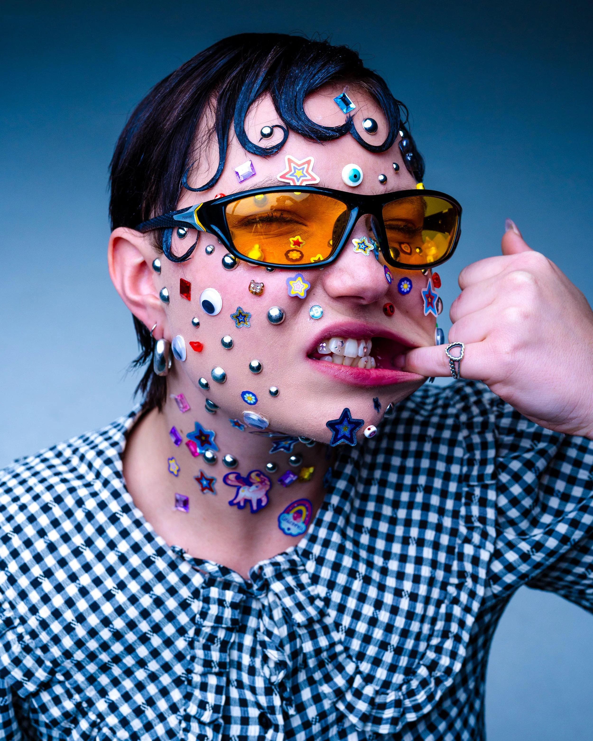 Person wearing yellow-tinted sunglasses, covered in colorful stickers, biting their finger, and wearing a black and white checkered shirt.