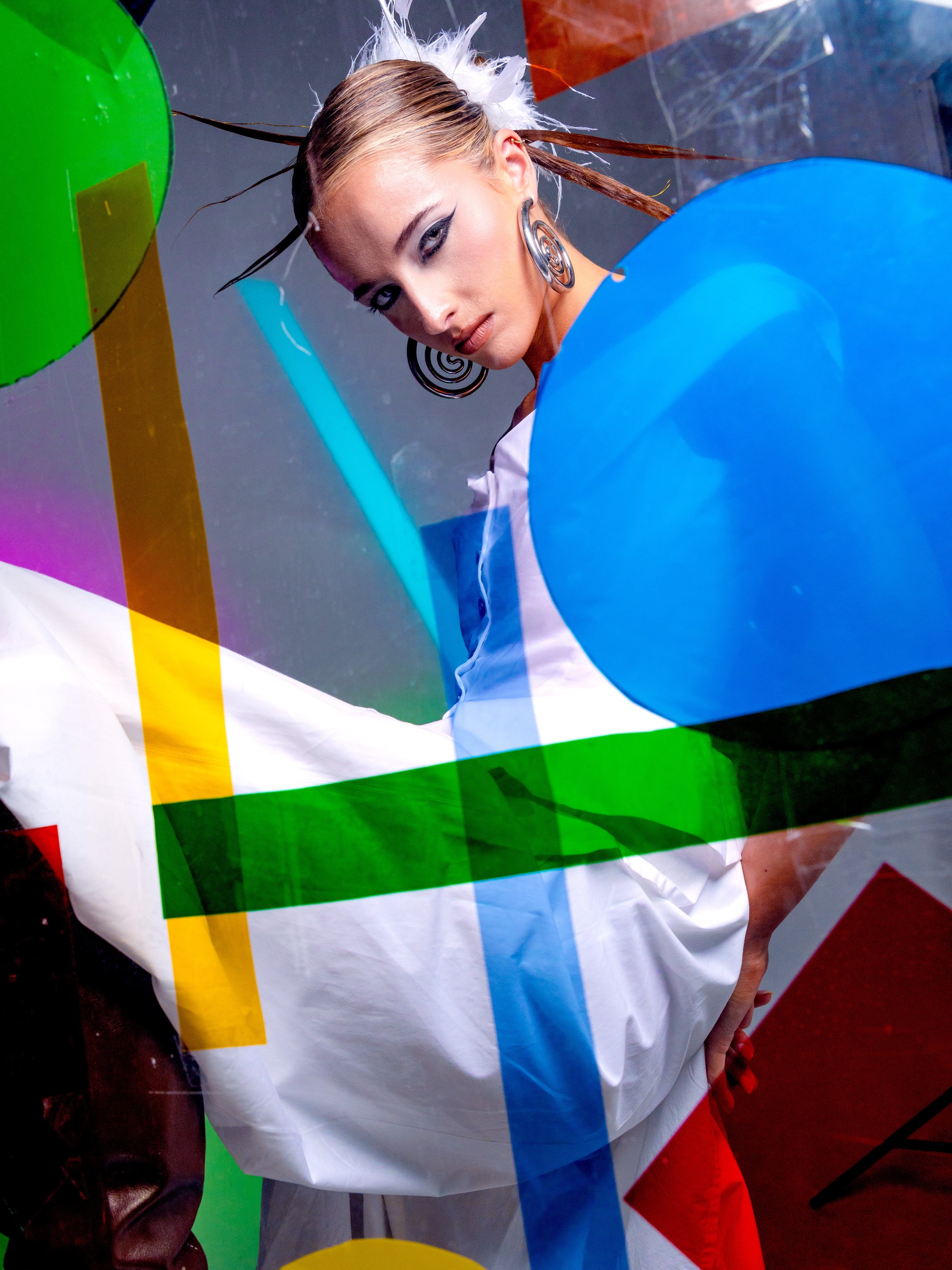 A woman with styled hair and bold makeup, wearing large spiral earrings, posing behind colorful abstract geometric shapes in a studio setting.