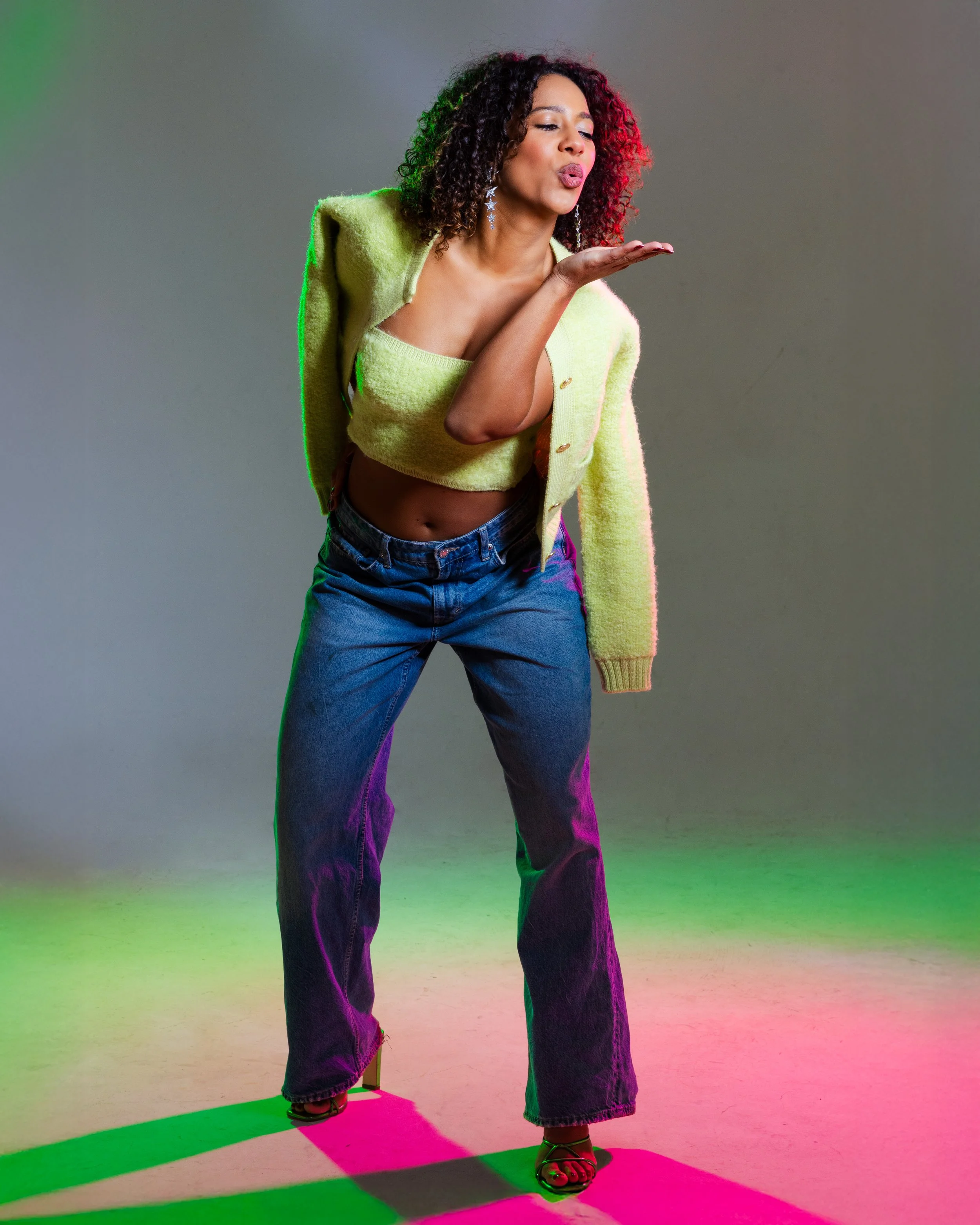 A woman with curly hair blowing a kiss, wearing a yellow cropped sweater with a matching cardigan, blue jeans, and heels, standing against a plain background with colored lighting.