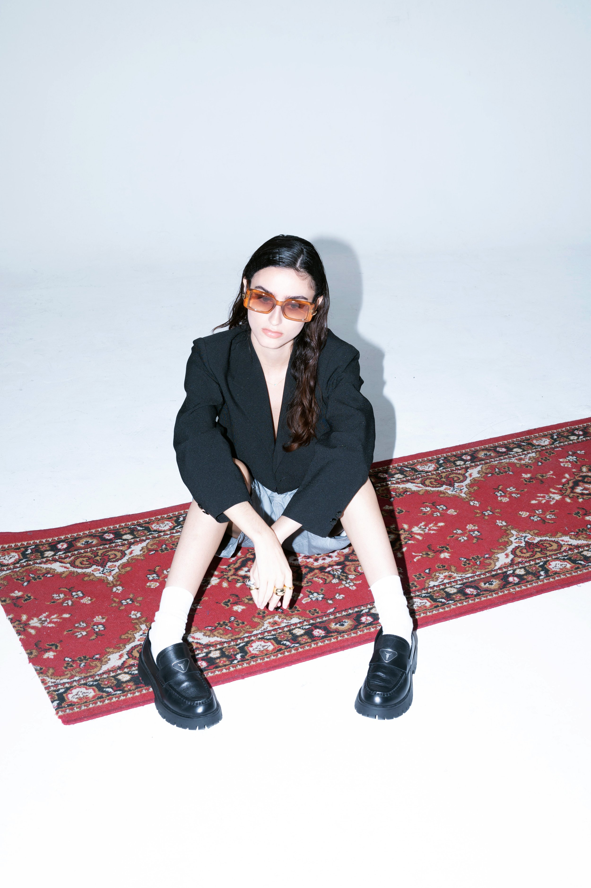 A woman with long dark hair wearing orange sunglasses, a black blazer, and white socks with black loafers, sitting on a red patterned rug in front of a plain white background.