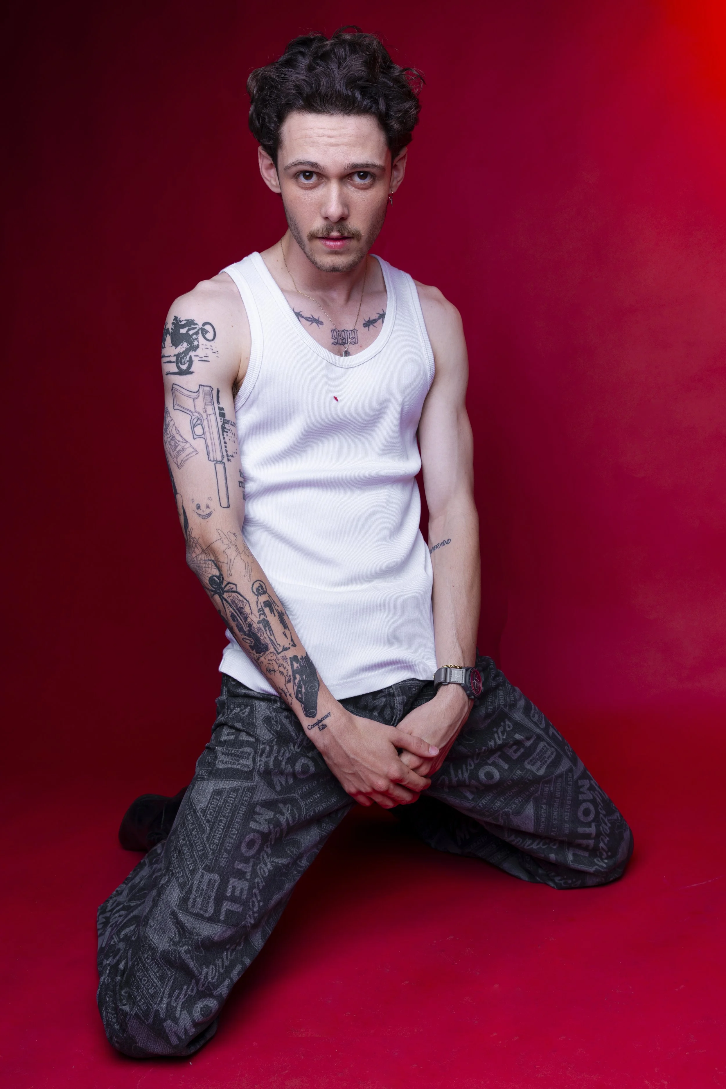 A young man with curly dark hair, wearing a white tank top and black patterned pants, kneeling on a red floor against a red background, displaying tattoos on his arms and chest.