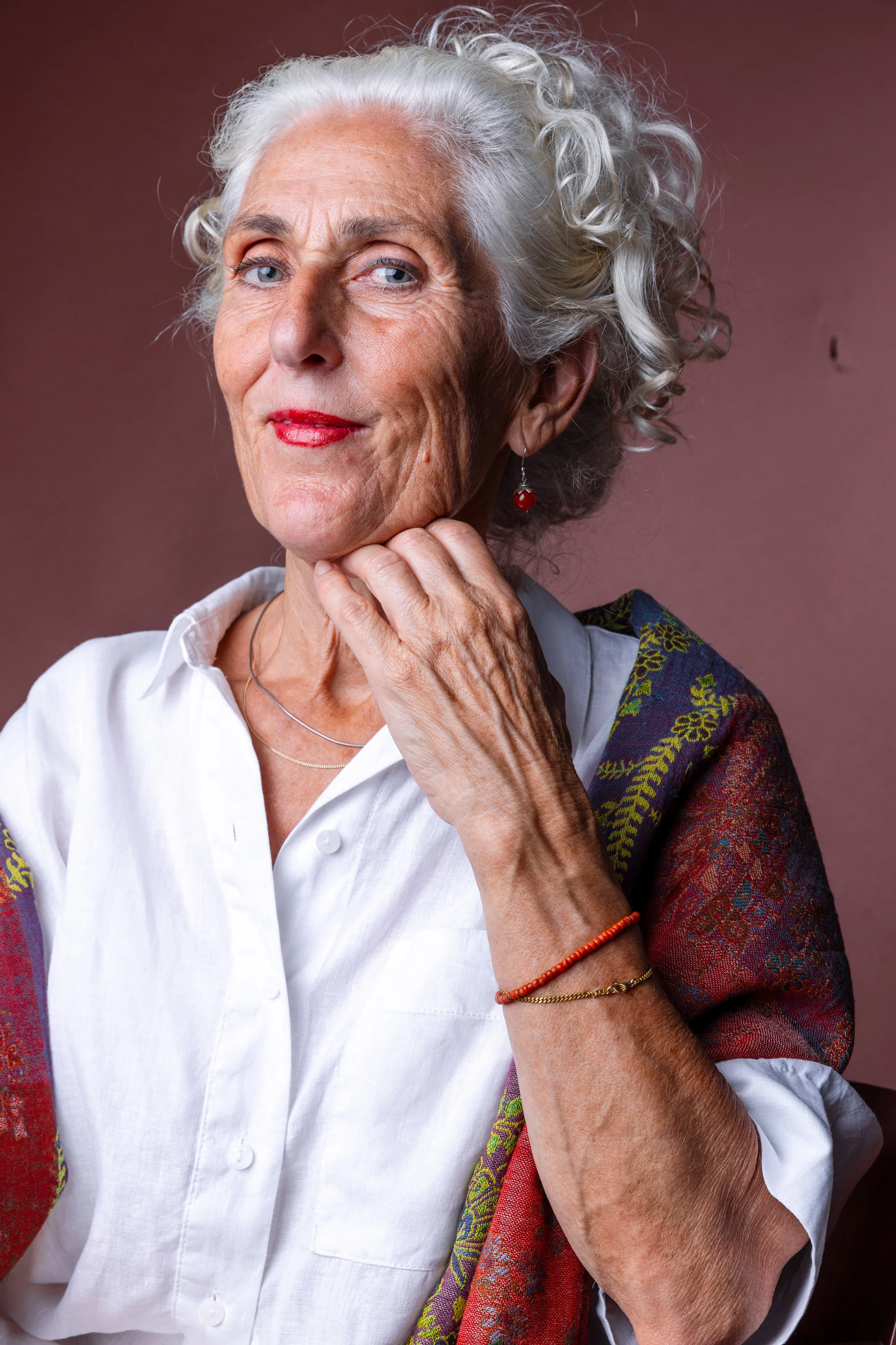 Portrait of an elderly woman with curly white hair, wearing a white shirt, red lipstick, and red earrings, resting her chin on her hand against a pink background.
