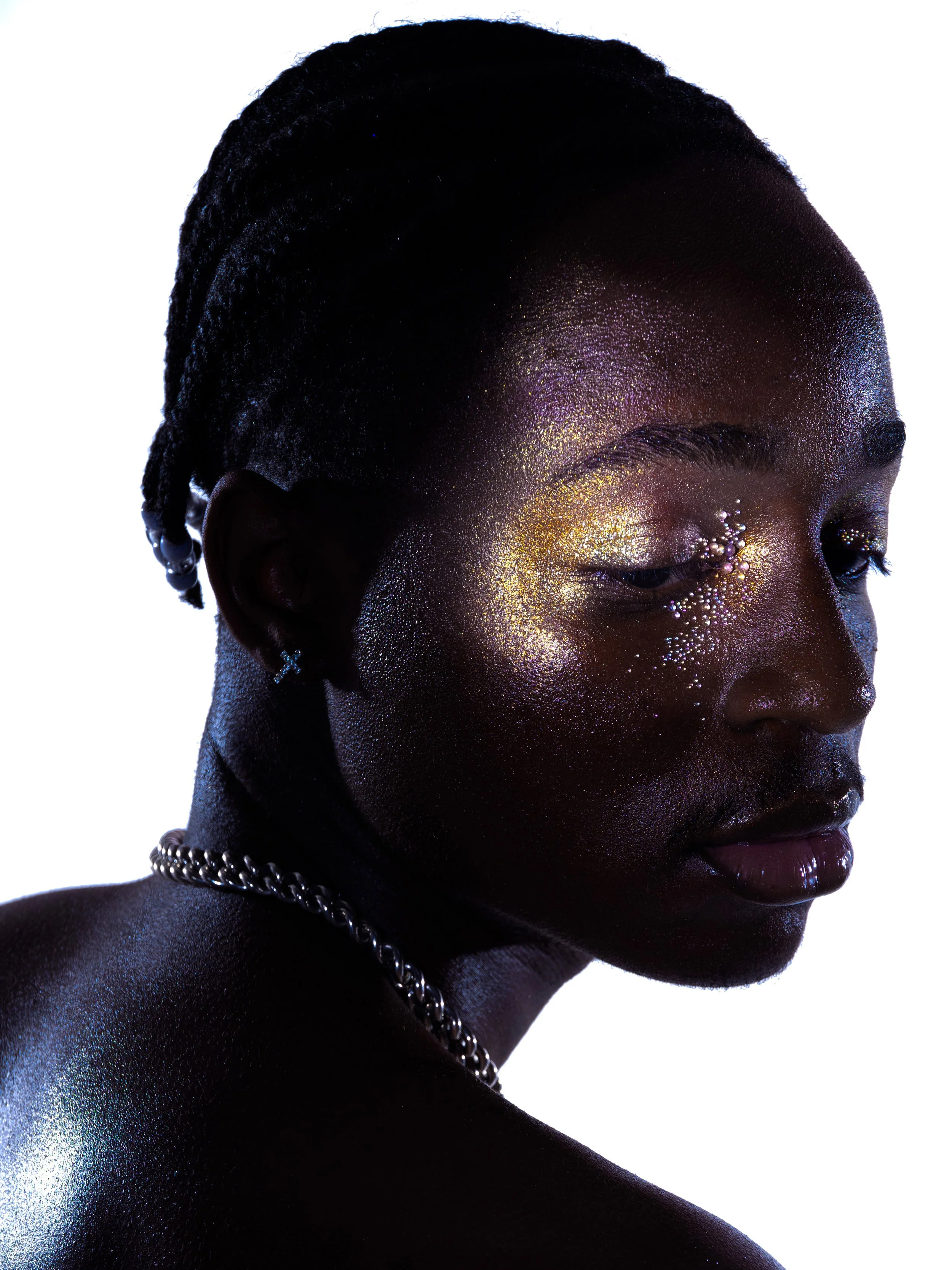 Close-up of a woman with dark skin and short braided hair, adorned with shimmering, iridescent makeup and glitter, against a white background.