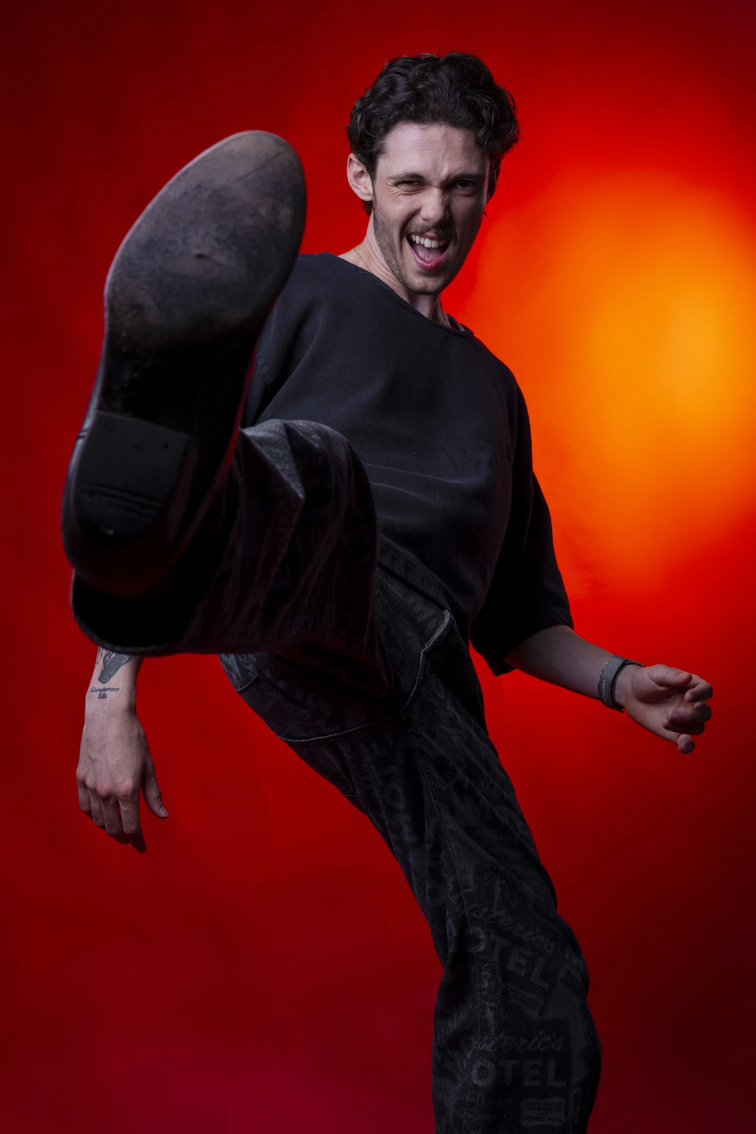 Young man in black clothing performing a high kick towards the camera, with a red background, smiling and winking.