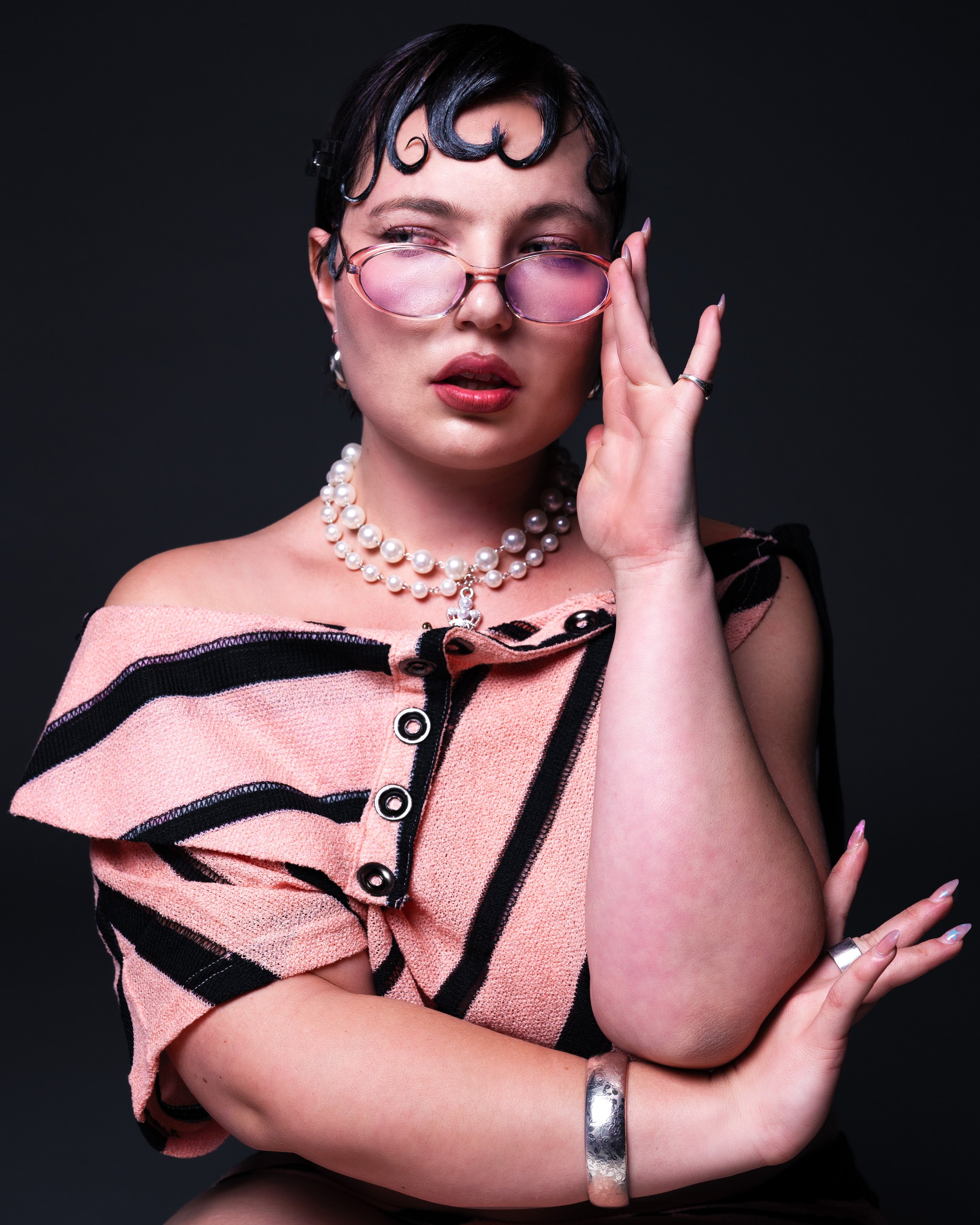 A woman with styled short black hair, pink tinted glasses, and vintage-inspired makeup, wearing a pink and black striped off-shoulder top, pearl necklaces, and jewelry, posing against a dark background.