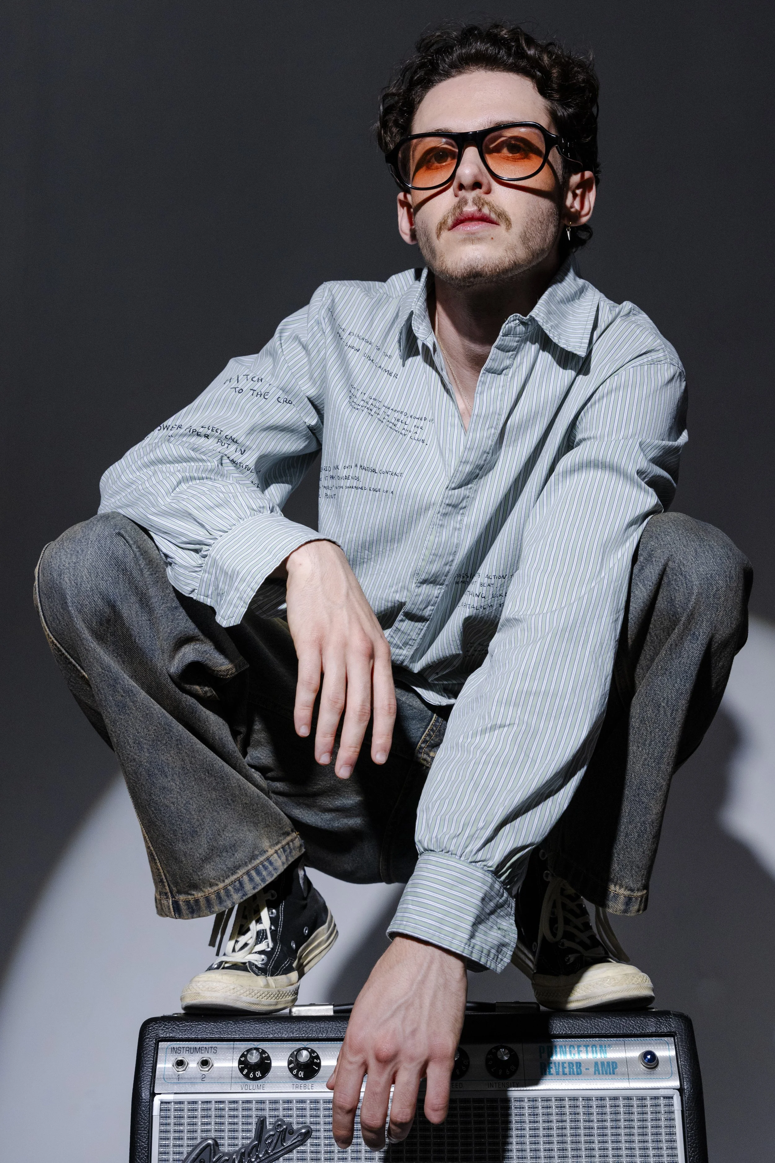 A young man with curly hair, wearing orange-tinted glasses, a striped shirt with writing, and gray jeans, squats on top of a Fender amplifier against a dark background.