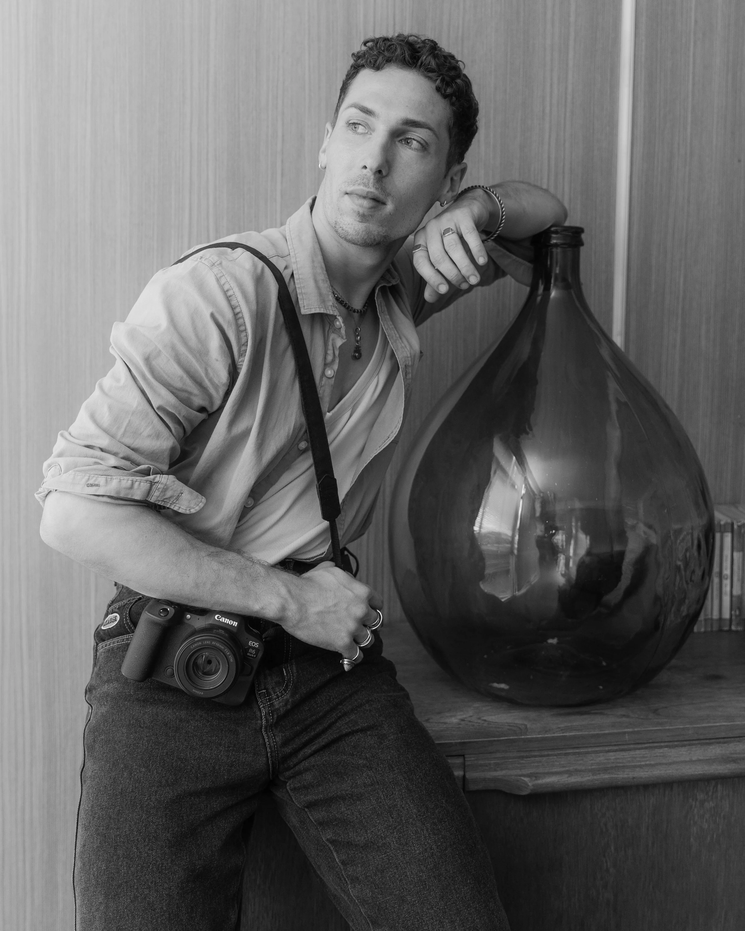 A young man with curly hair leaning on a large glass vase with his left arm, holding a camera with his right hand, standing in front of a wooden wall.