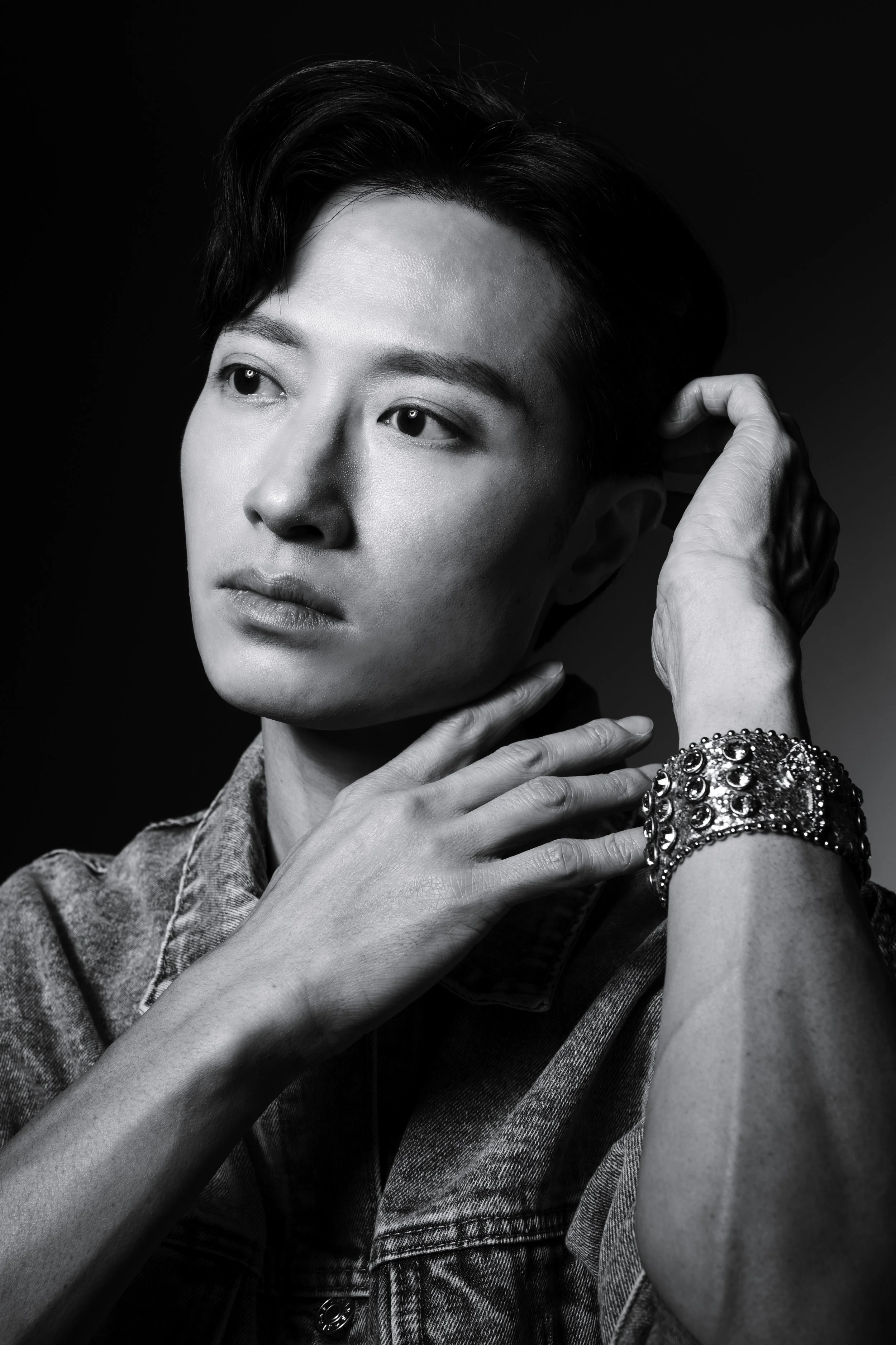 Black and white portrait of a young person with short dark hair, wearing a denim jacket and a large, ornate bracelet, looking contemplative with hands near face.