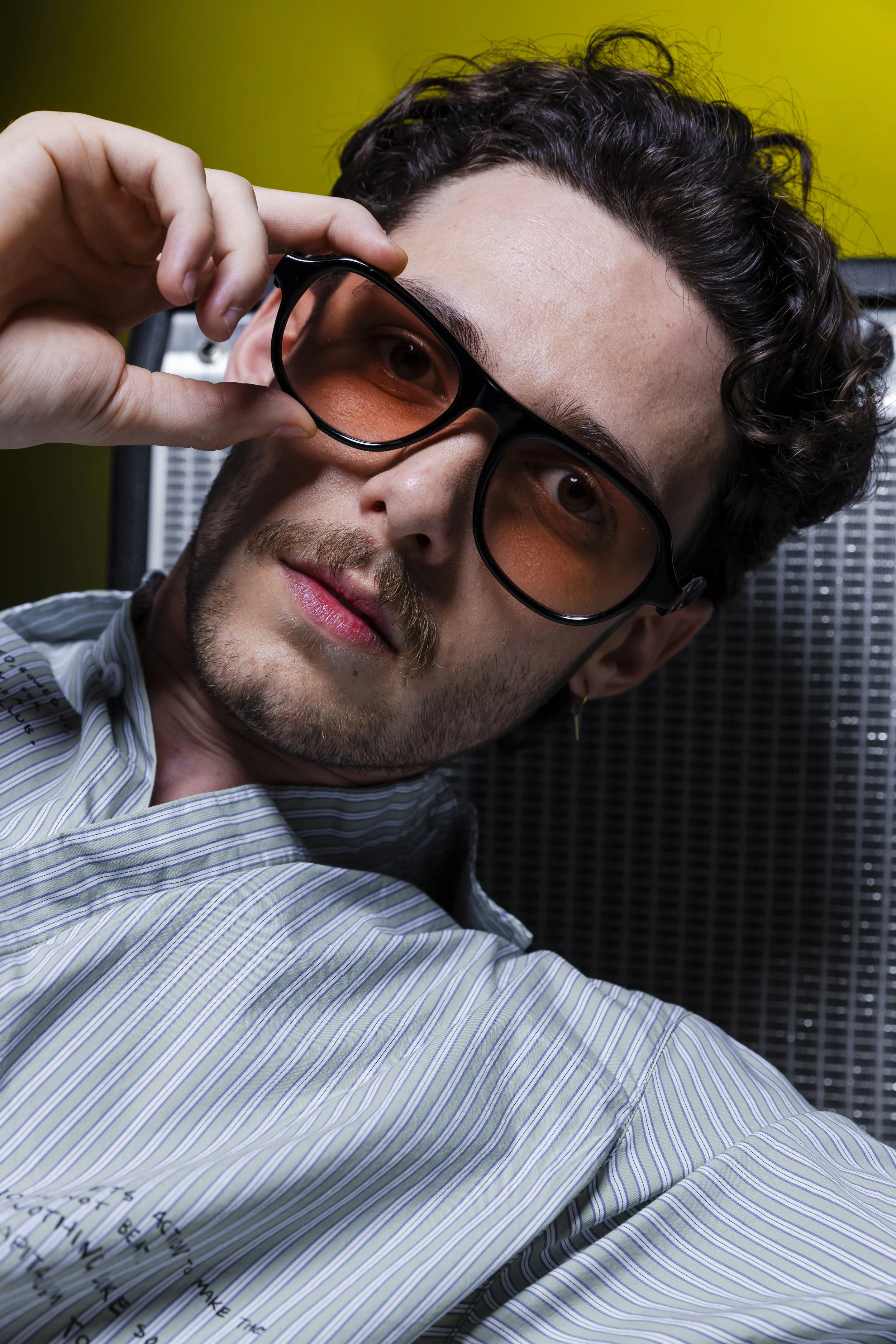 A young man with curly dark hair, wearing striped shirt and sunglasses, is adjusting his glasses and looking into the camera with a serious expression, against a greenish-yellow background.