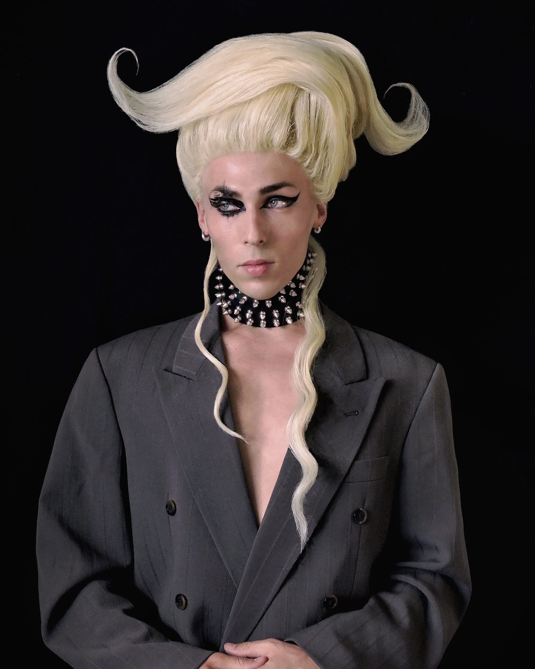 Woman with elaborate platinum blonde hair styled in dramatic, swirling waves, wearing a dark pinstripe suit, choker with spikes, and black makeup around one eye, against a black background.