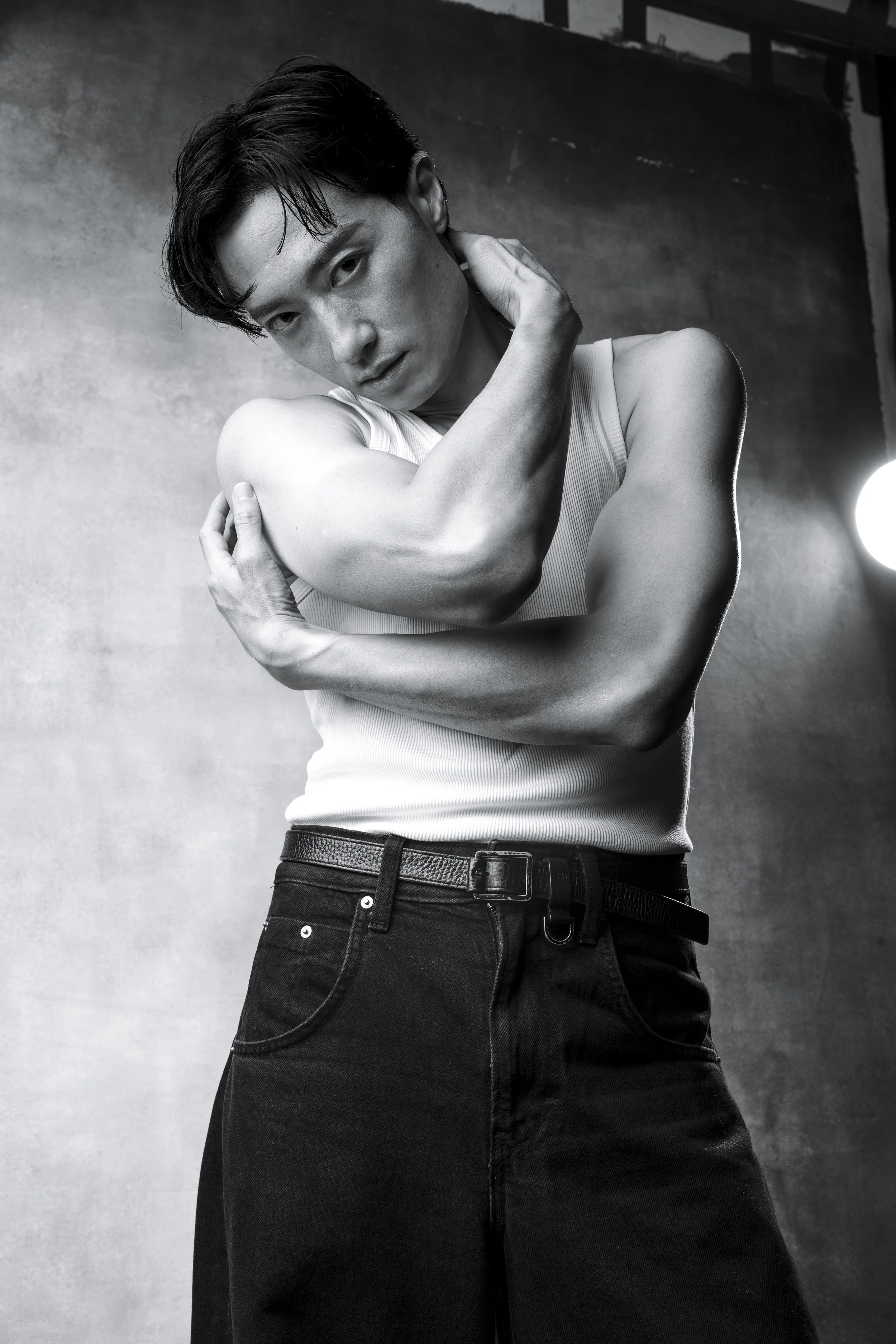 A black and white photo of a young man with dark hair, wearing a sleeveless shirt and black jeans, posing with his arms crossed over his chest and one hand touching his neck, looking directly at the camera with a serious expression.