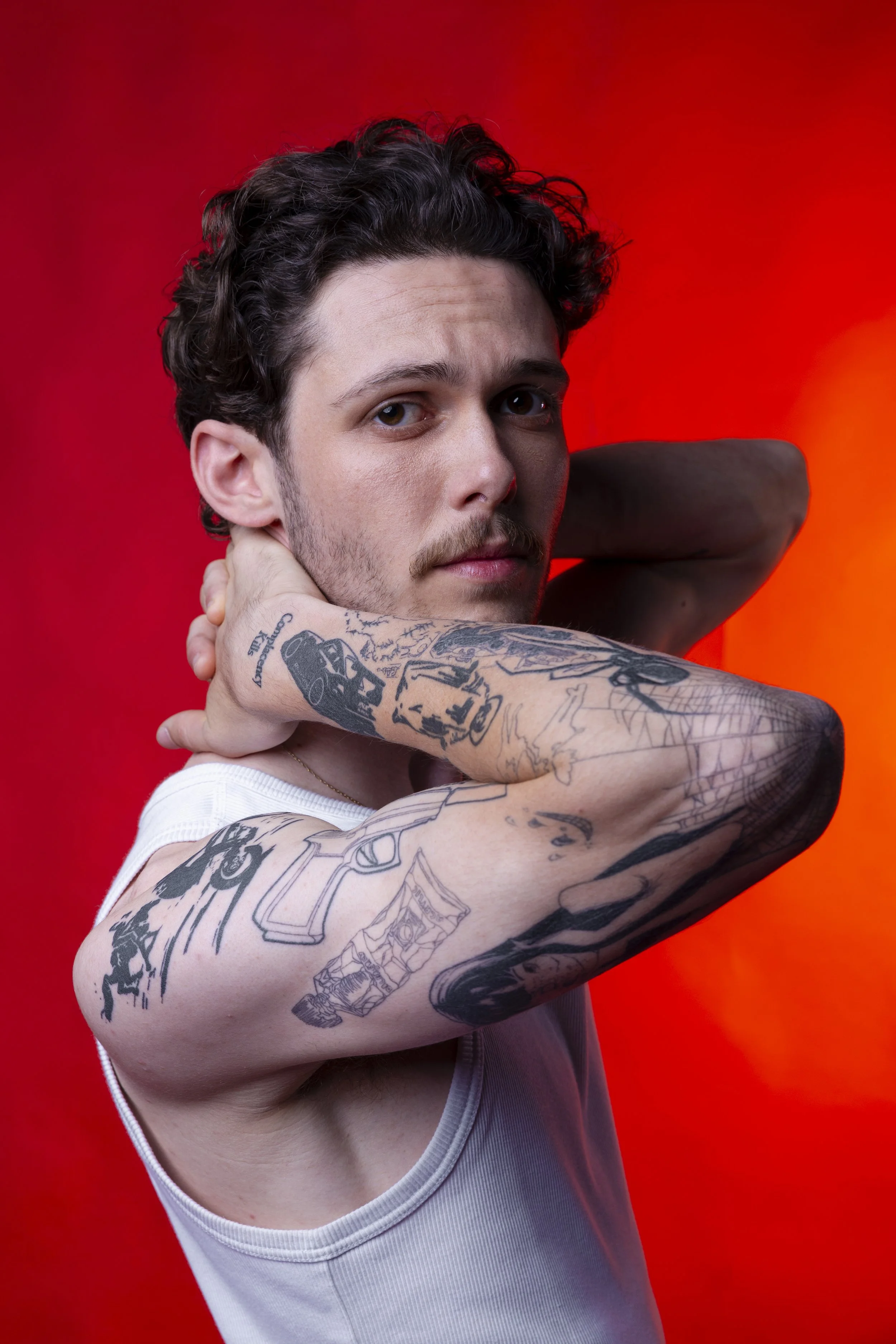 A young man with tattoos on his arms and chest, posing with one arm bent and his hand behind his head, against a red background.