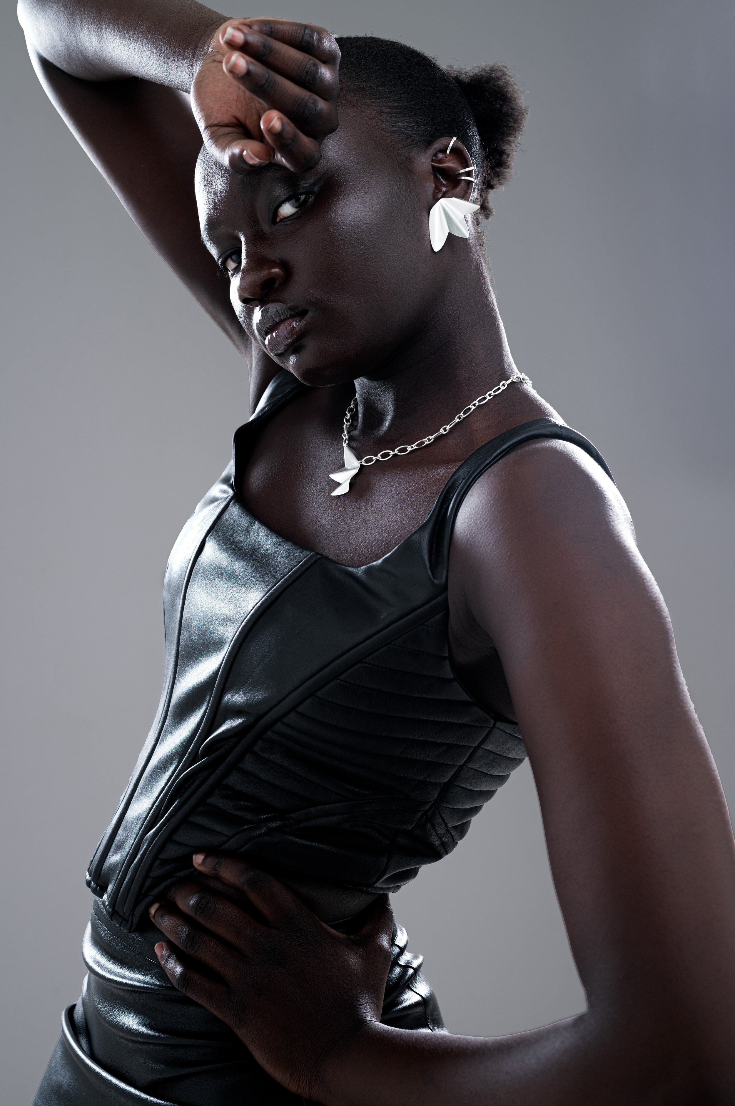 A woman with dark skin and natural hair styled in a short puff, wearing a black leather sleeveless top and matching pants. She accessorizes with a silver chain necklace with a bird charm, white earrings, and multiple ear cuffs. She poses with one arm