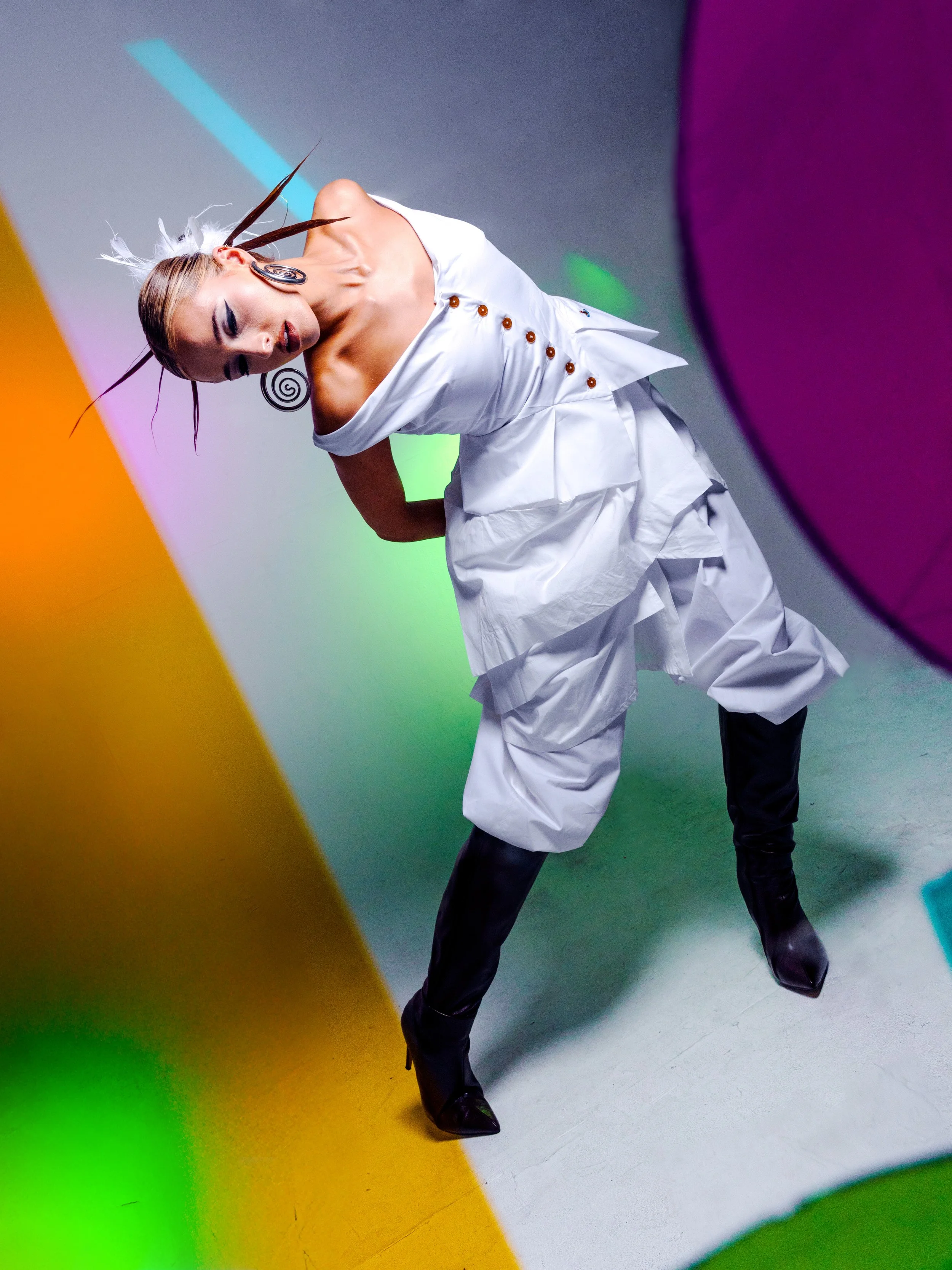 A woman dressed in a futuristic white outfit with black boots and unique makeup poses against a colorful abstract background with yellow, purple, green, and pink elements.