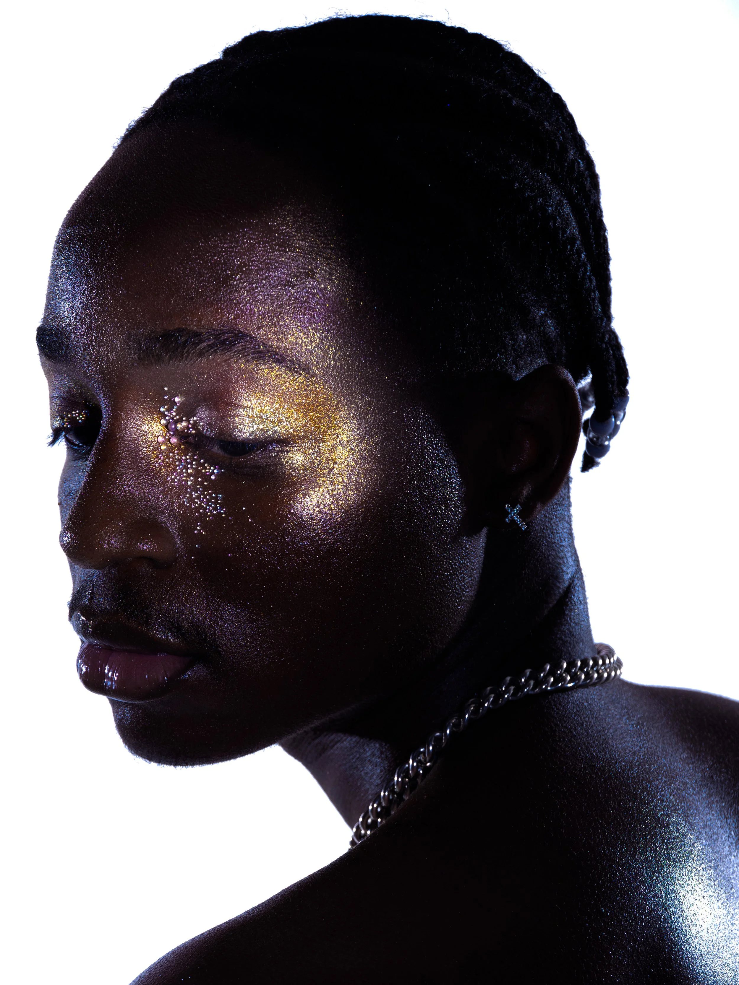 Close-up of a woman with dark skin and short hair, illuminated with iridescent makeup and glitter, wearing earrings and a chain necklace against a white background.