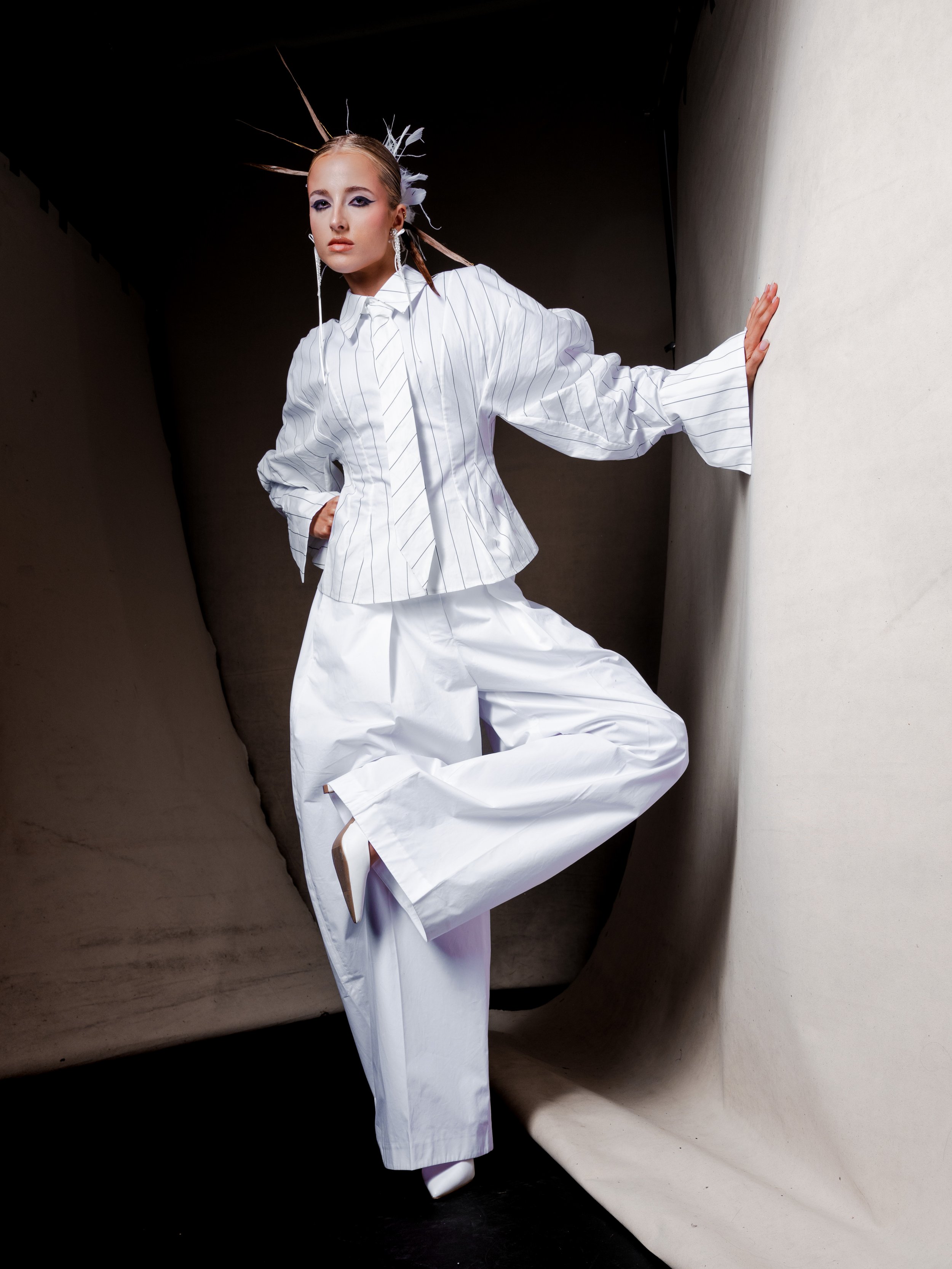 Fashion model wearing a white pinstripe top and wide-leg pants, striking a pose against a creative curved backdrop with dramatic lighting.