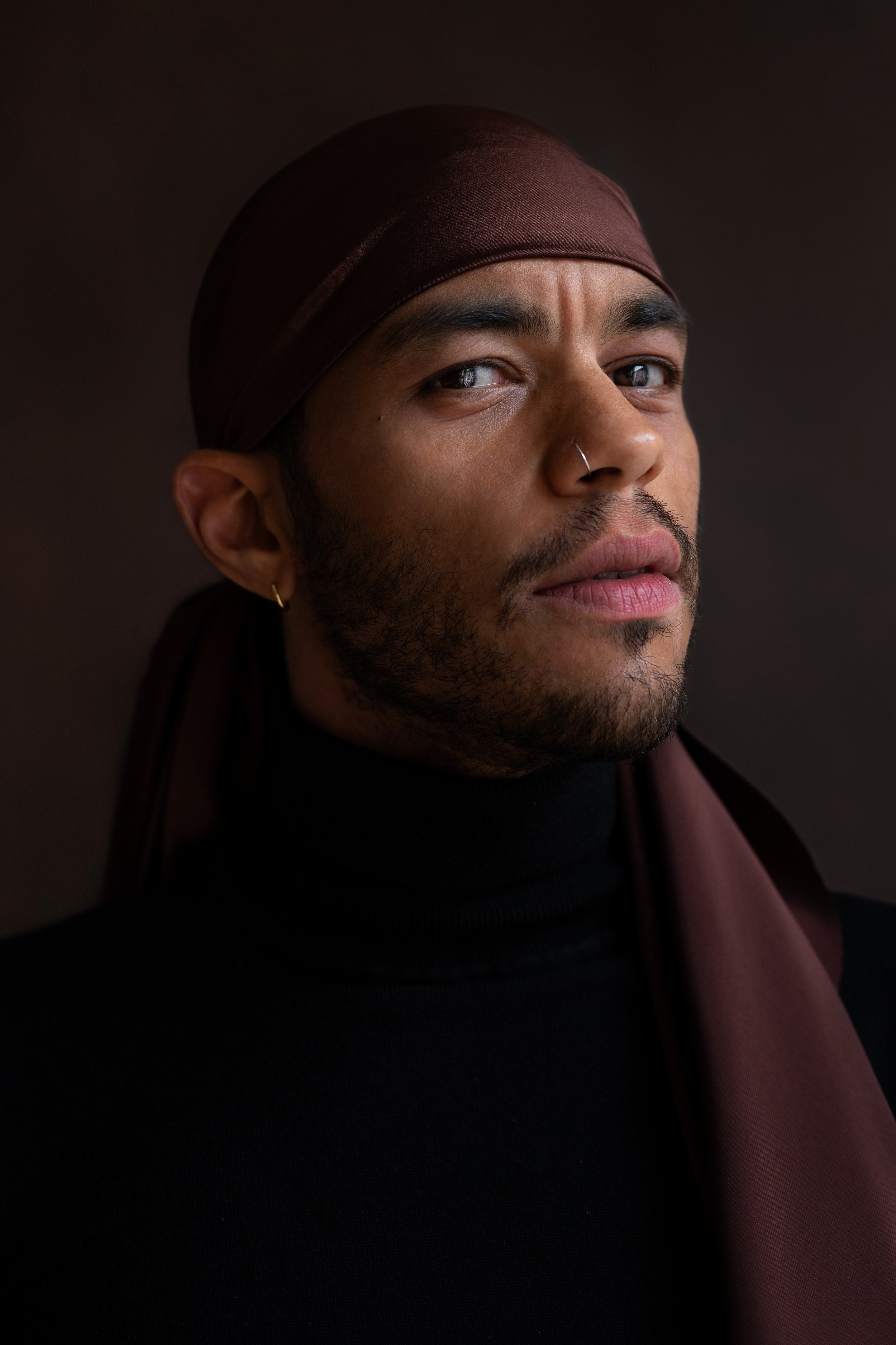 Portrait of a man wearing a dark brown head wrap and black turtleneck, with earrings and a nose ring, looking slightly to the side with a confident expression.
