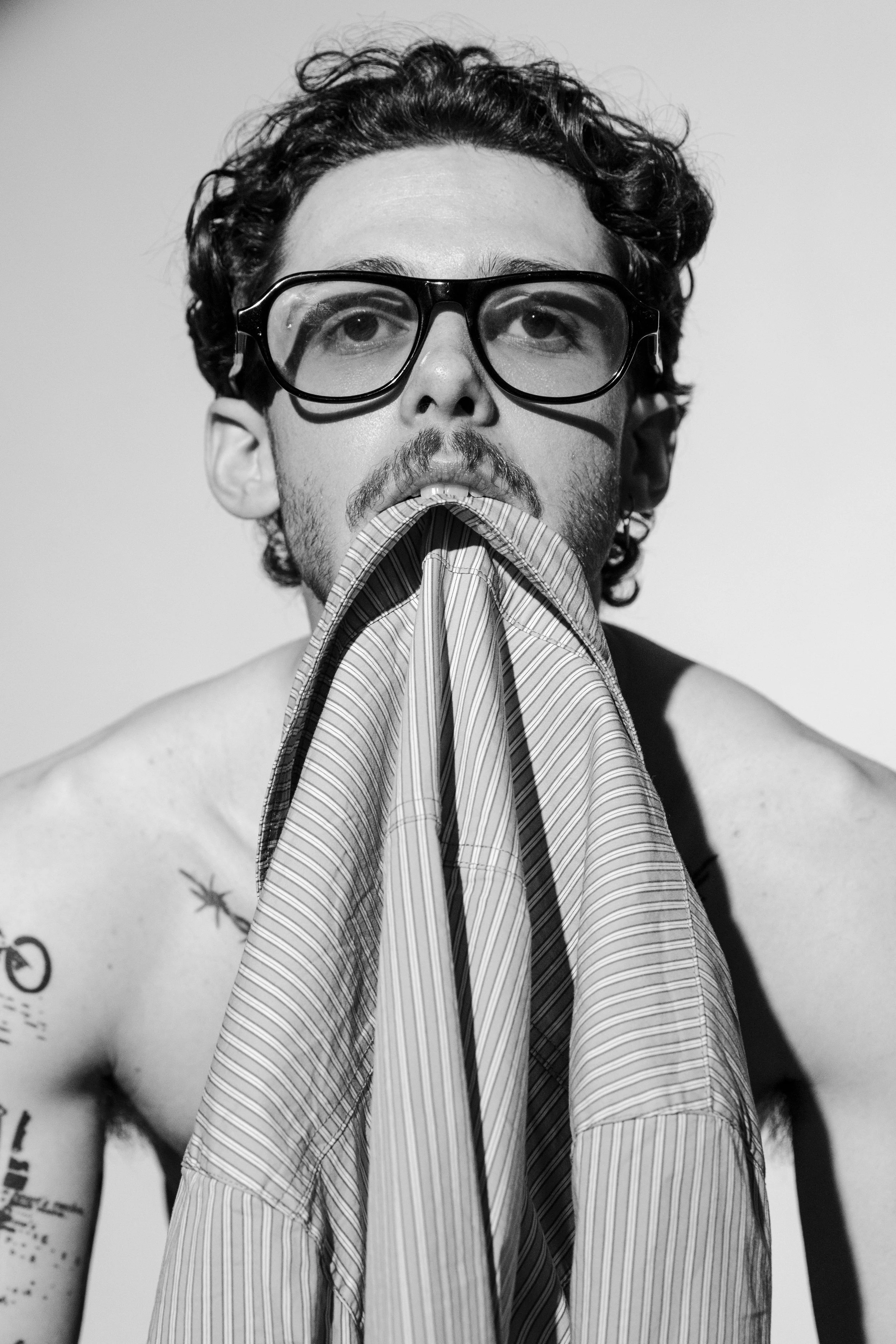 A man with curly hair and glasses is biting down on a striped dress shirt, pulling it out of his mouth. He is shirtless, showing tattoos on his left shoulder and arm, and has a background with a plain wall.