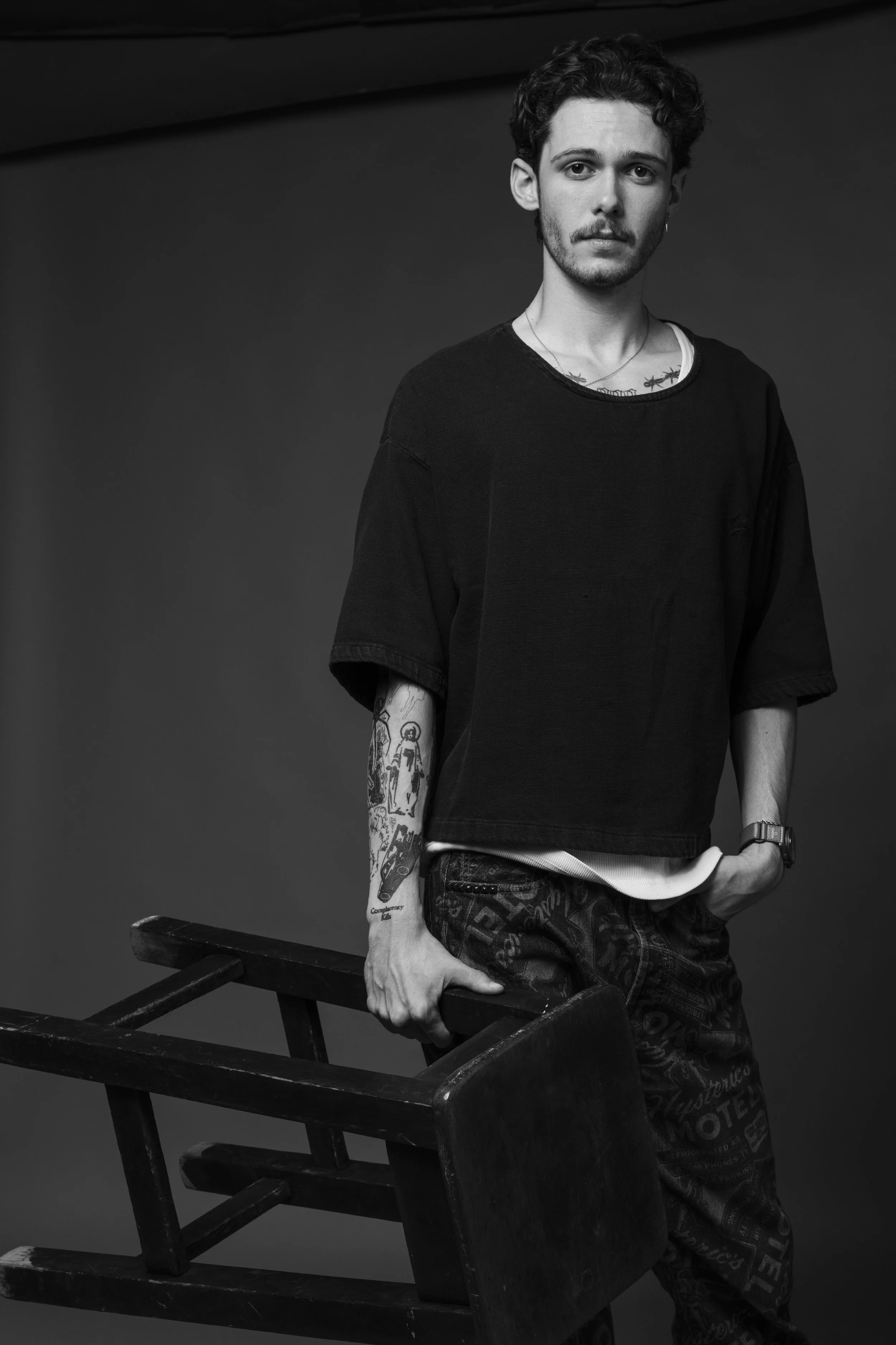 Black and white photo of a young man with curly hair and tattoos wearing a loose black T-shirt and patterned pants, holding a wooden chair in a studio setting.