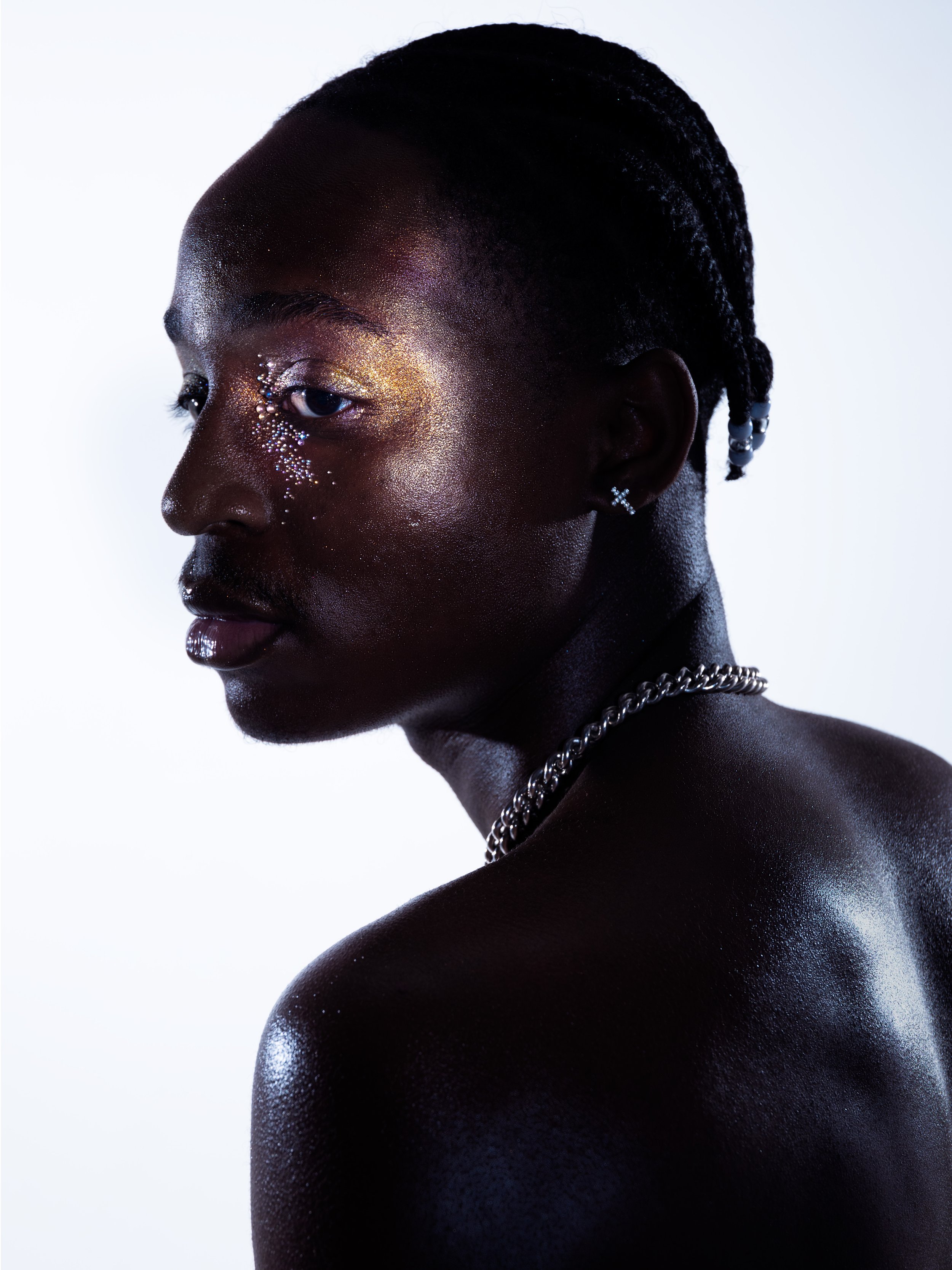 A close-up profile of a dark-skinned person with short, textured hair and makeup with glitter and rhinestones around one eye, wearing earrings, a silver chain necklace, and with bare shoulders against a white background.
