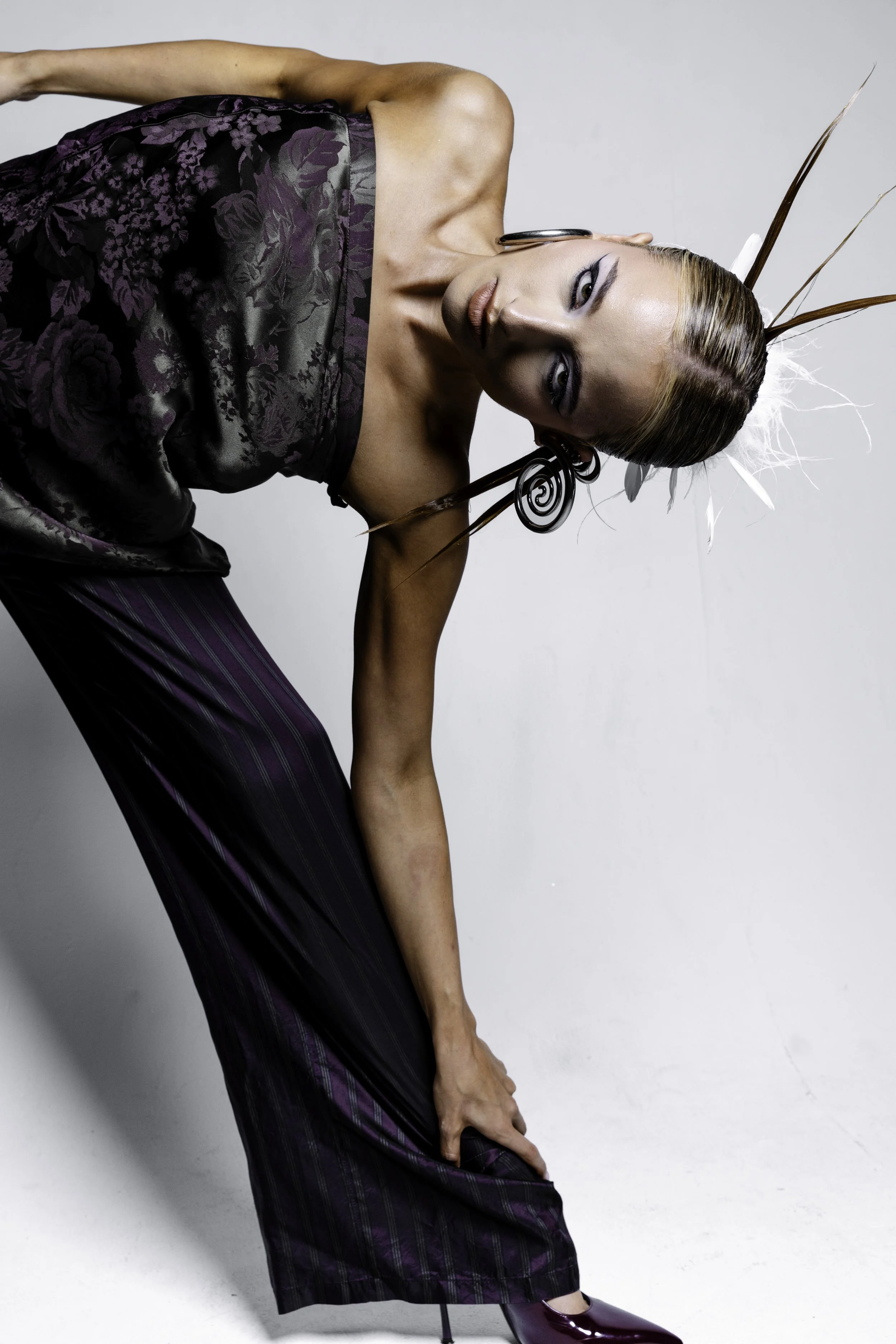 A woman dressed in an elaborate outfit with feathers and jewelry, bending forward with one hand on her leg against a plain background.