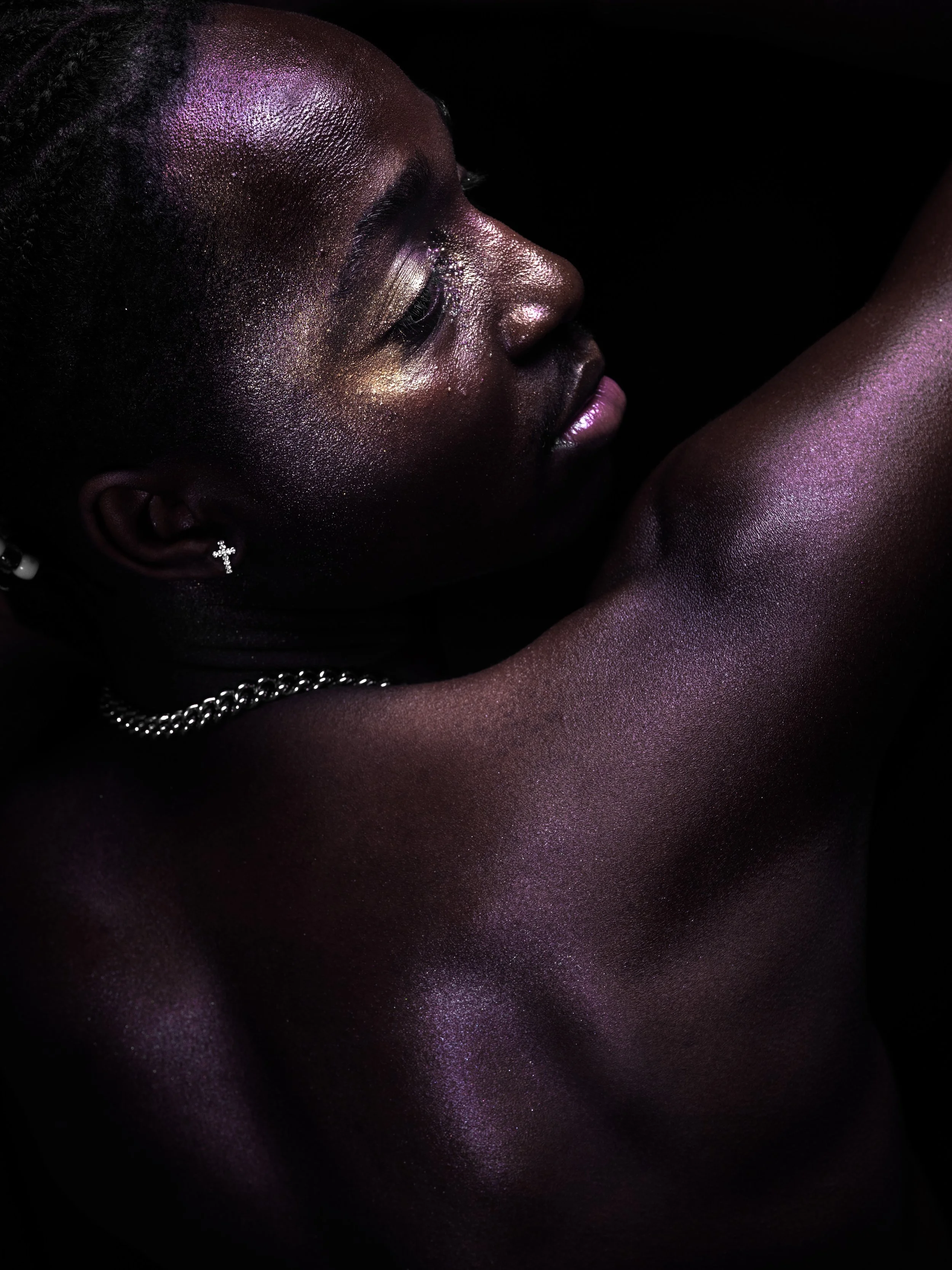Close-up side profile of a woman with dark skin, glittery makeup, and jewelry, with her arm raised against a black background