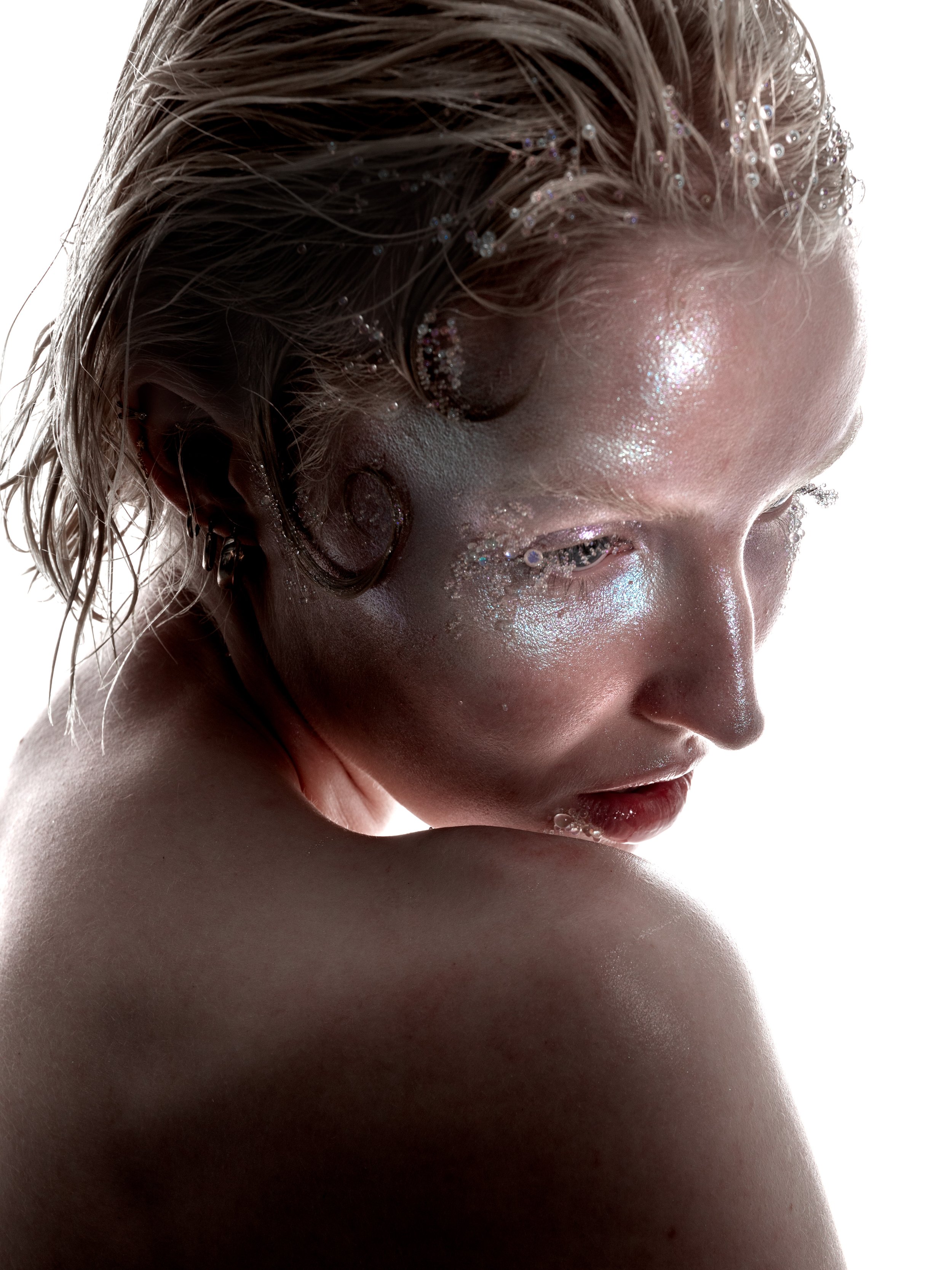 Close-up of a woman with glitter on her face, wet-looking hair, and a contemplative expression, looking downward over her shoulder.