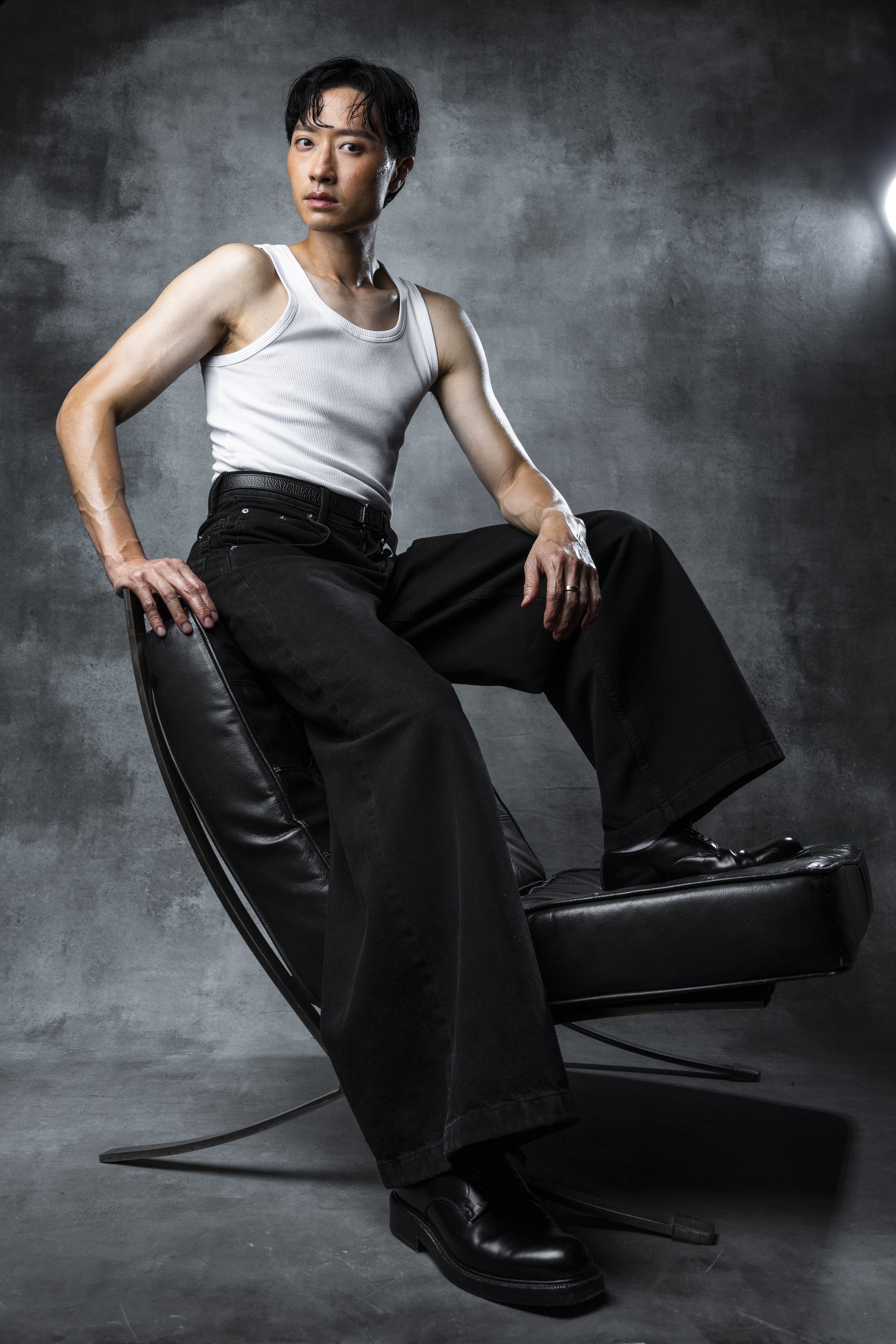 A person with short black hair, wearing a white tank top, black wide-leg pants, and black shoes, sitting on a black leather chair against a gray textured background.