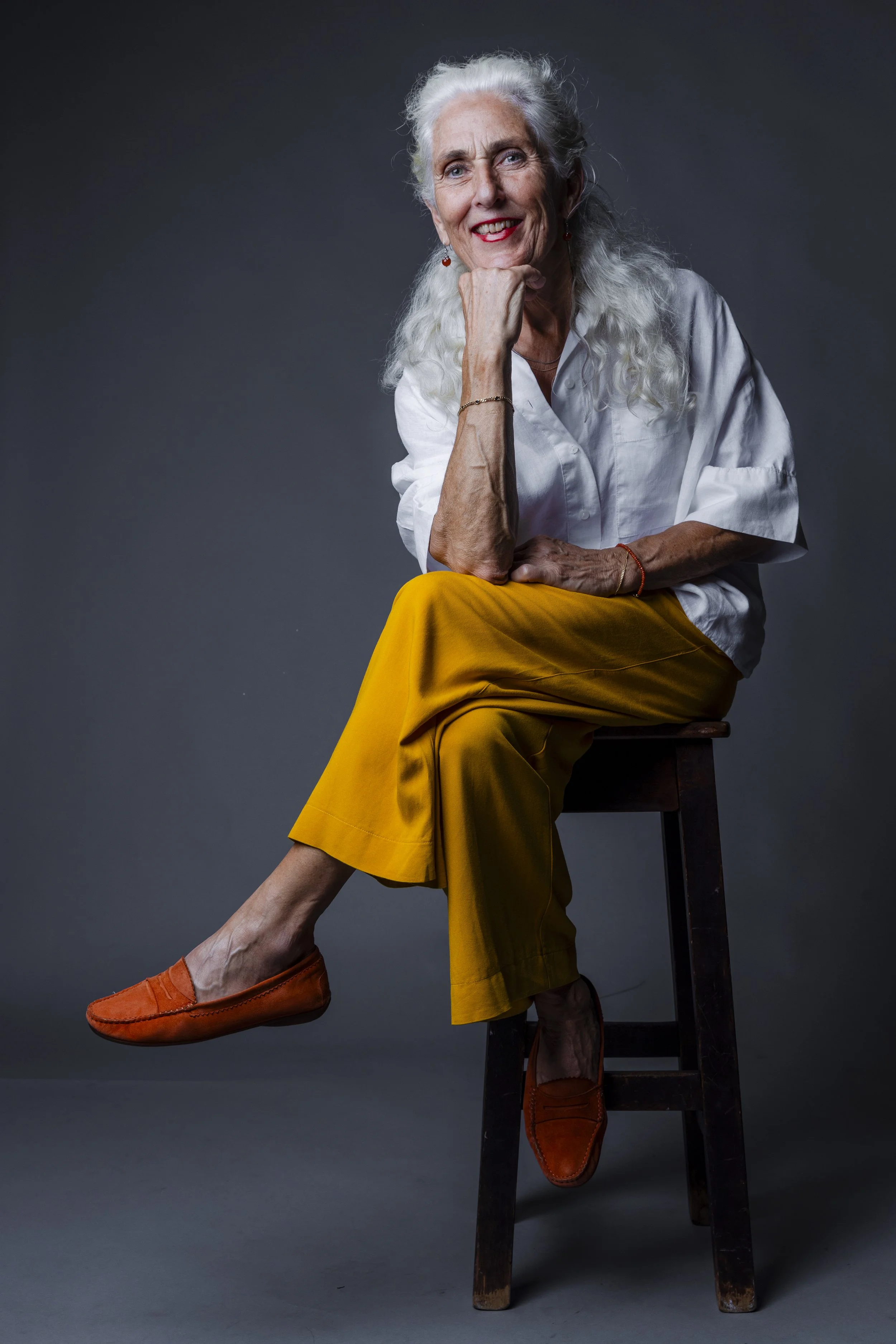 An elderly woman with white hair, wearing a white blouse and mustard yellow pants, sitting on a wooden stool against a dark background, smiling gently, with her chin resting on her hand.