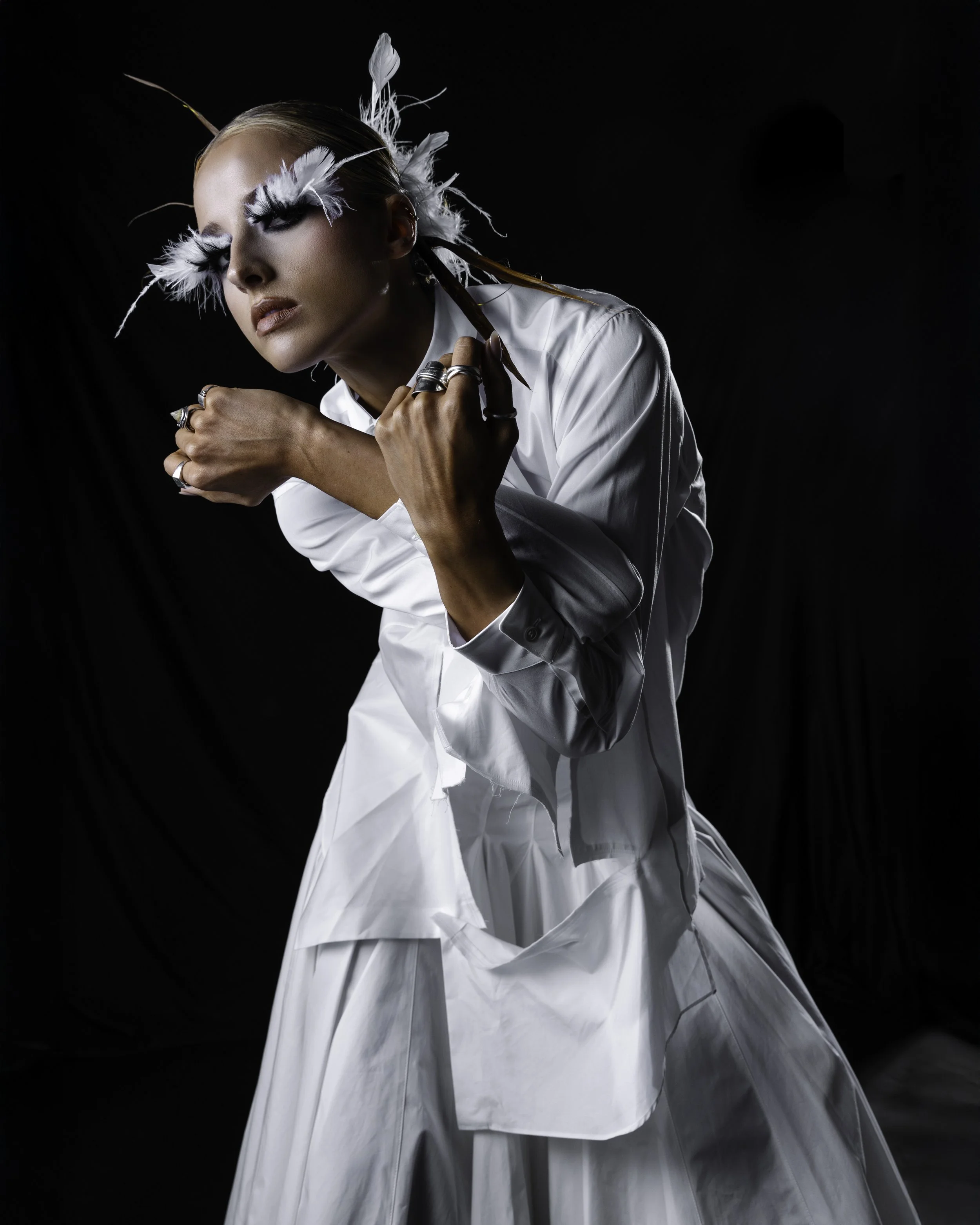 A woman with elaborate feathered eye makeup and jewelry, dressed in a white outfit, poses against a black background.