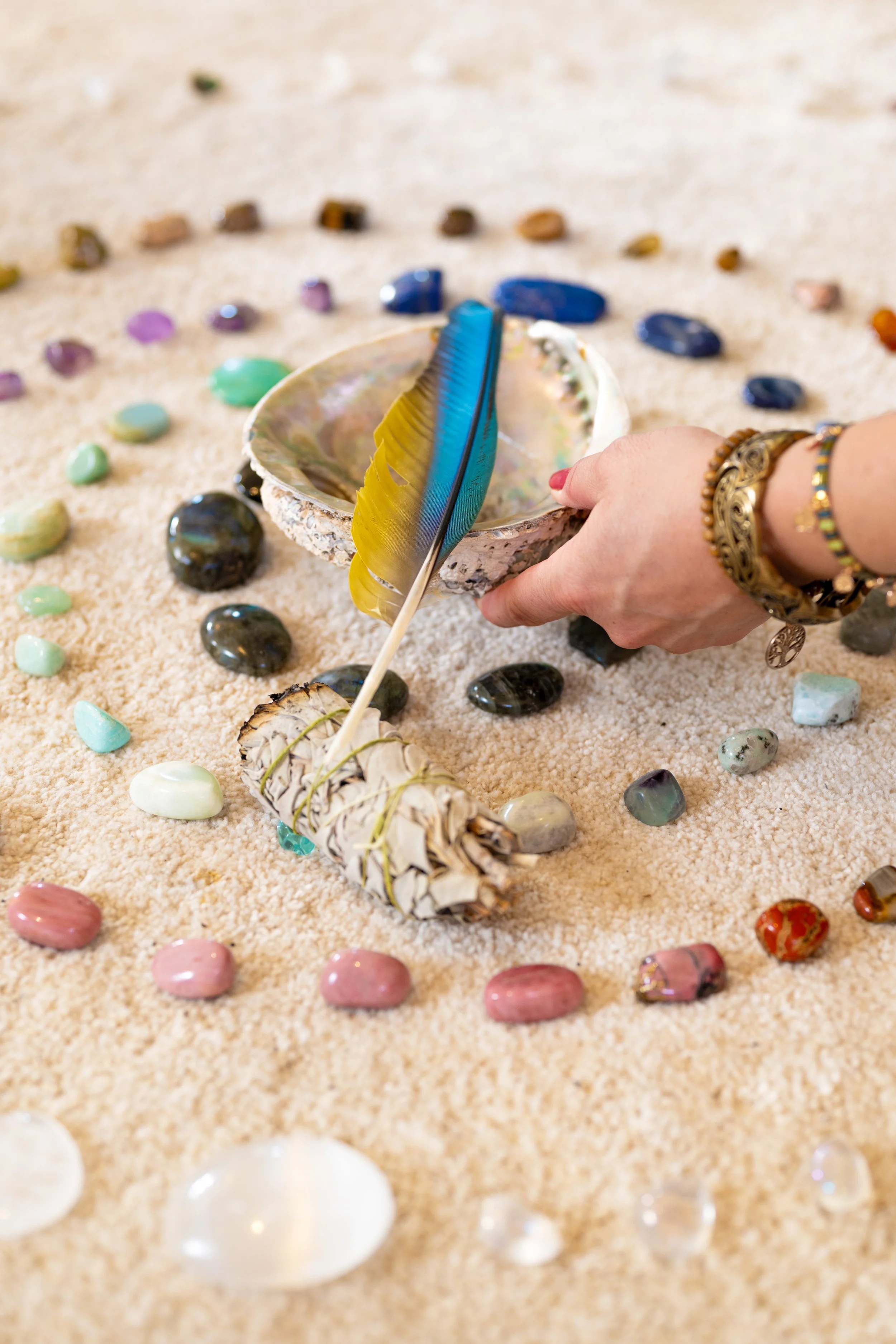 Person arranging colorful stones and seashells on sand with a shell and feather in center.