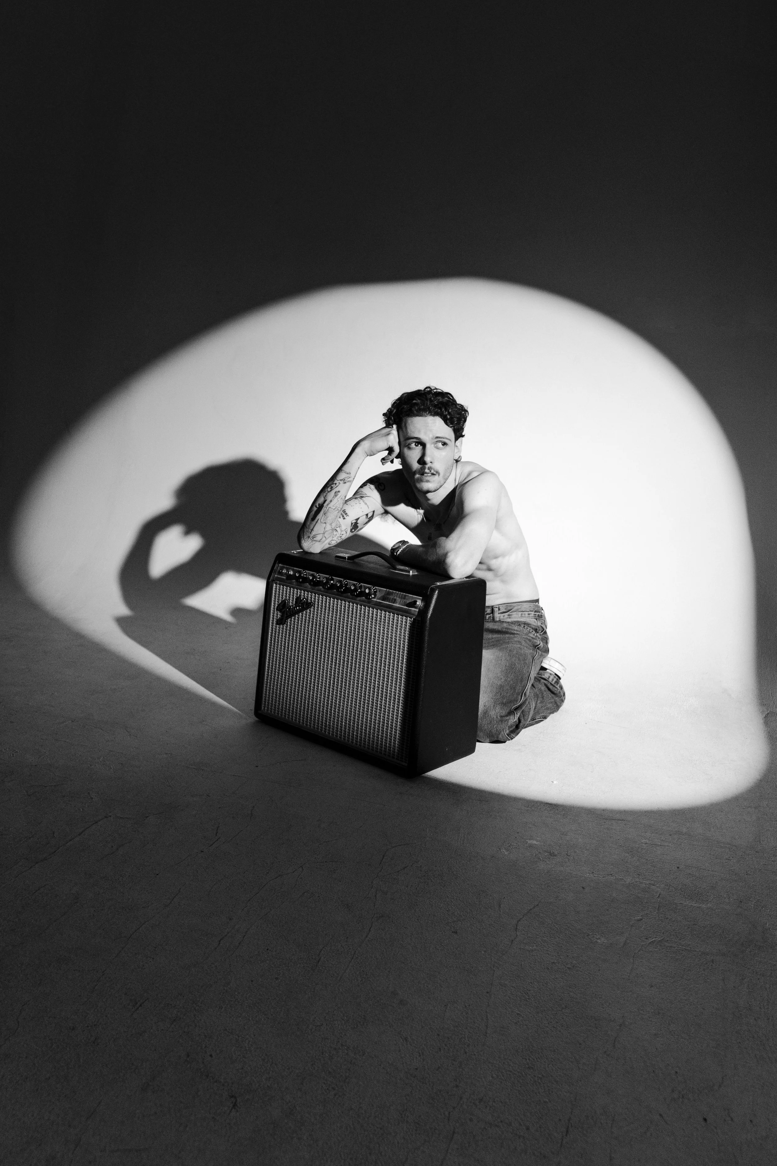 A black-and-white photo of a shirtless man with tattoos sitting on the floor, leaning on a guitar amplifier, under a spotlight creating a shadow on the wall.