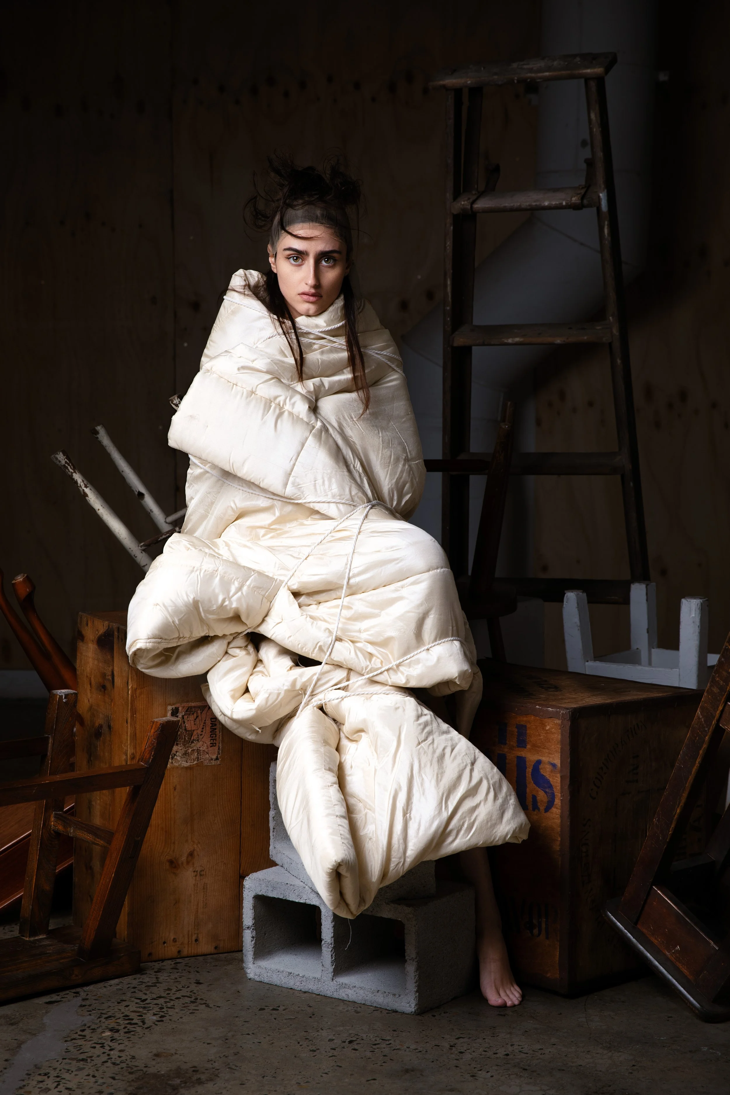 A woman sitting on concrete blocks, wrapped in a large cream-colored bag, surrounded by wooden chairs and a step ladder, in a dimly lit room with wooden walls.