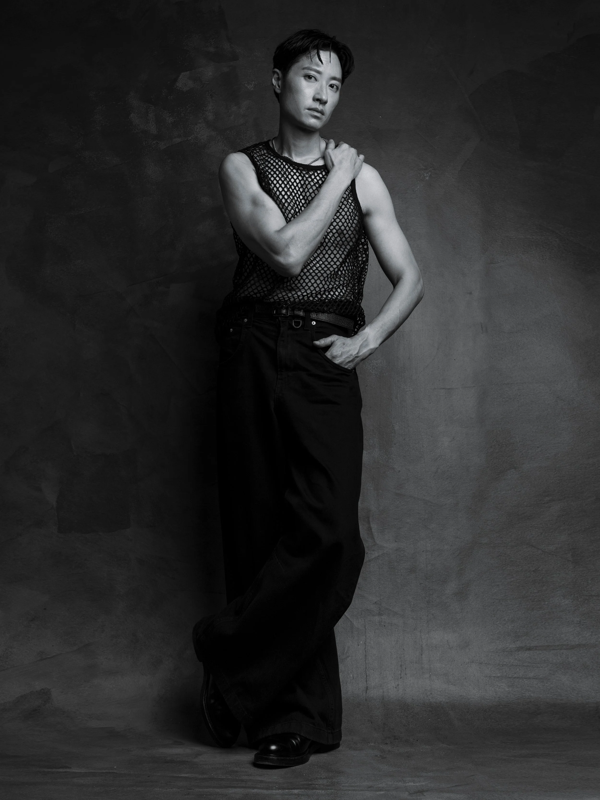 Black and white photo of a person posing against a textured wall, wearing a sleeveless mesh top and wide-leg pants, with short hair and a serious expression.