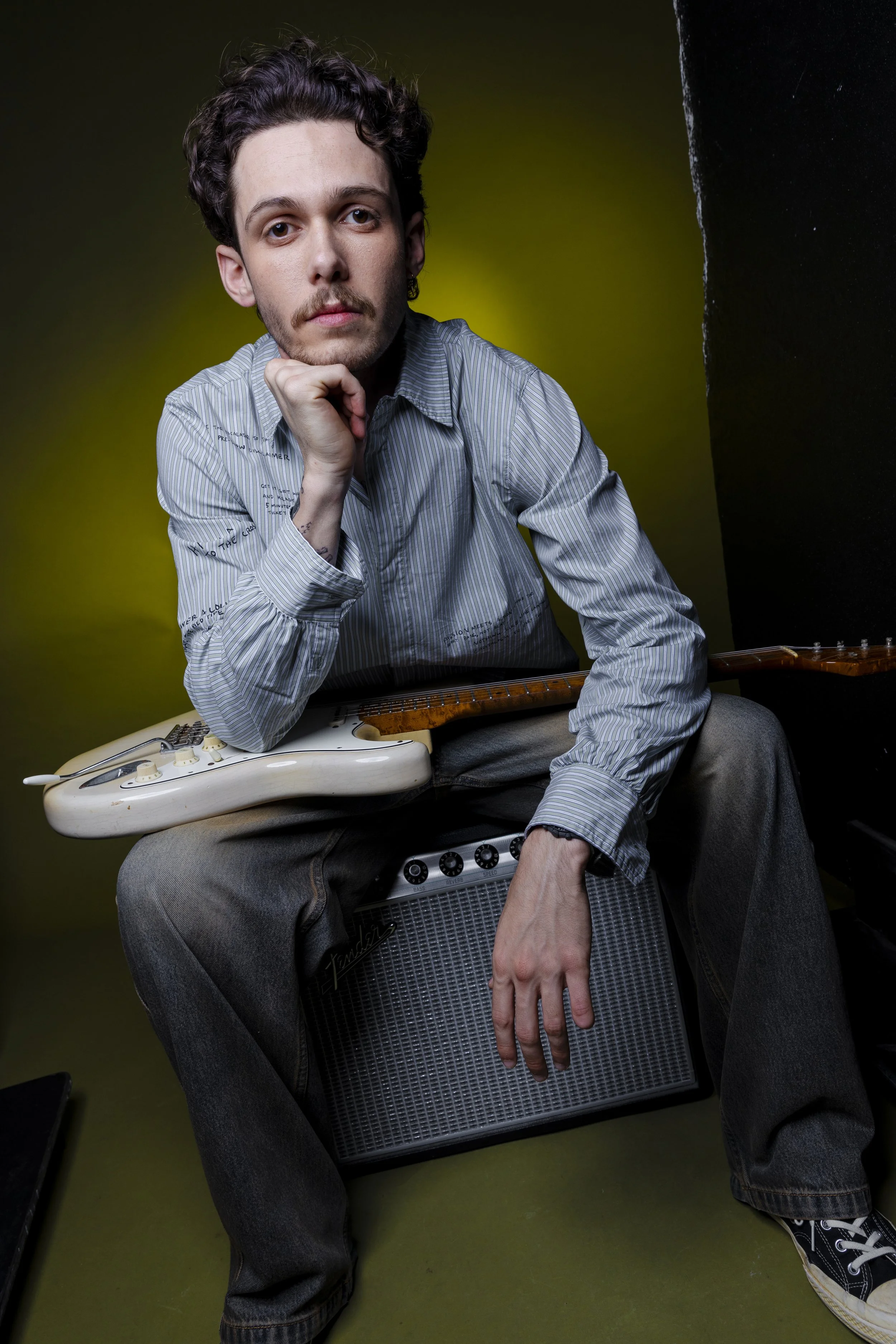 A young man with dark curly hair, wearing a striped shirt, sitting with an electric guitar resting on his lap, and a guitar amplifier beneath his hand, against a dark green and black background.