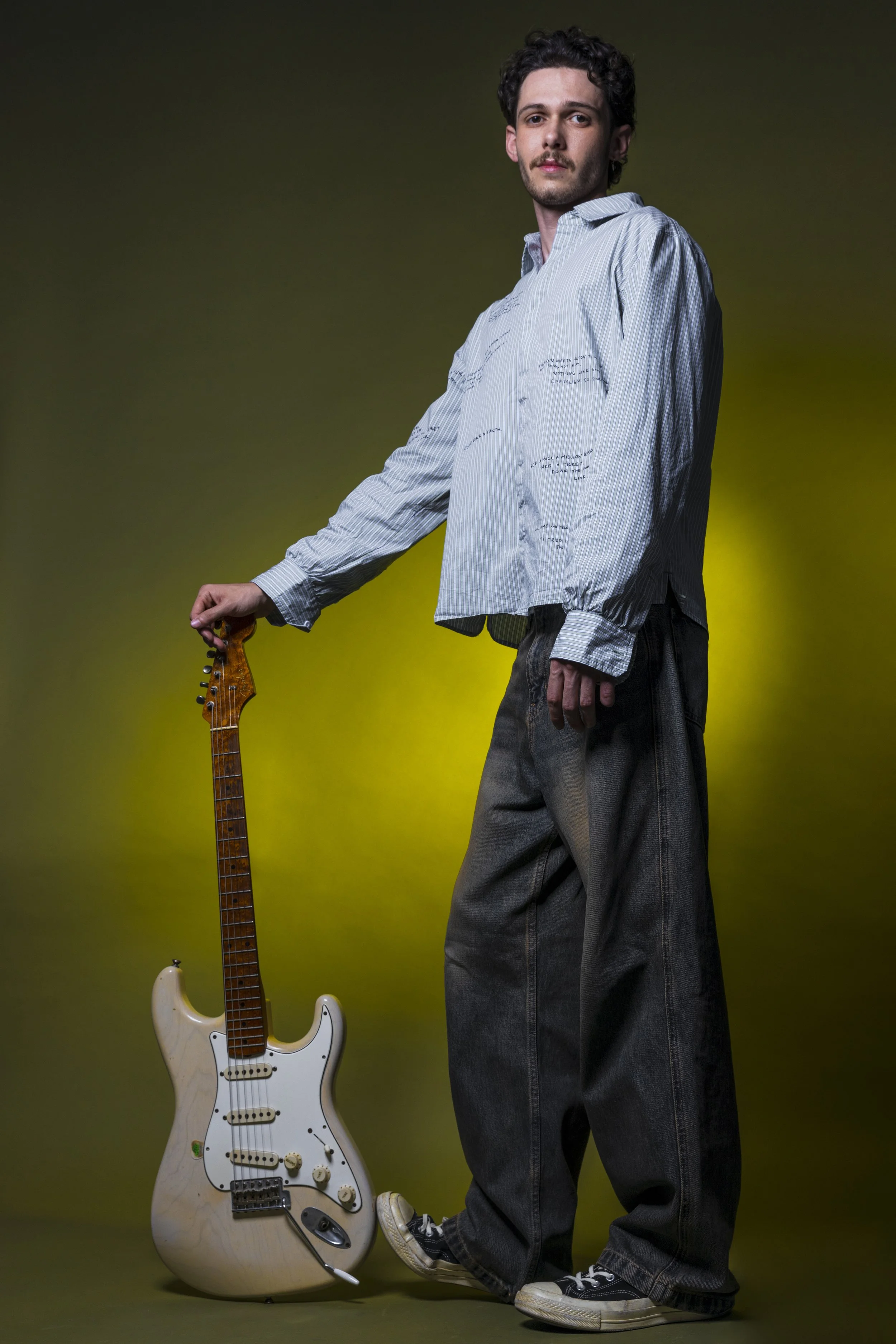 A young man with dark curly hair and a mustache standing against a gradient background, holding an electric guitar by his side, wearing a light-colored, long-sleeve shirt and baggy dark jeans.