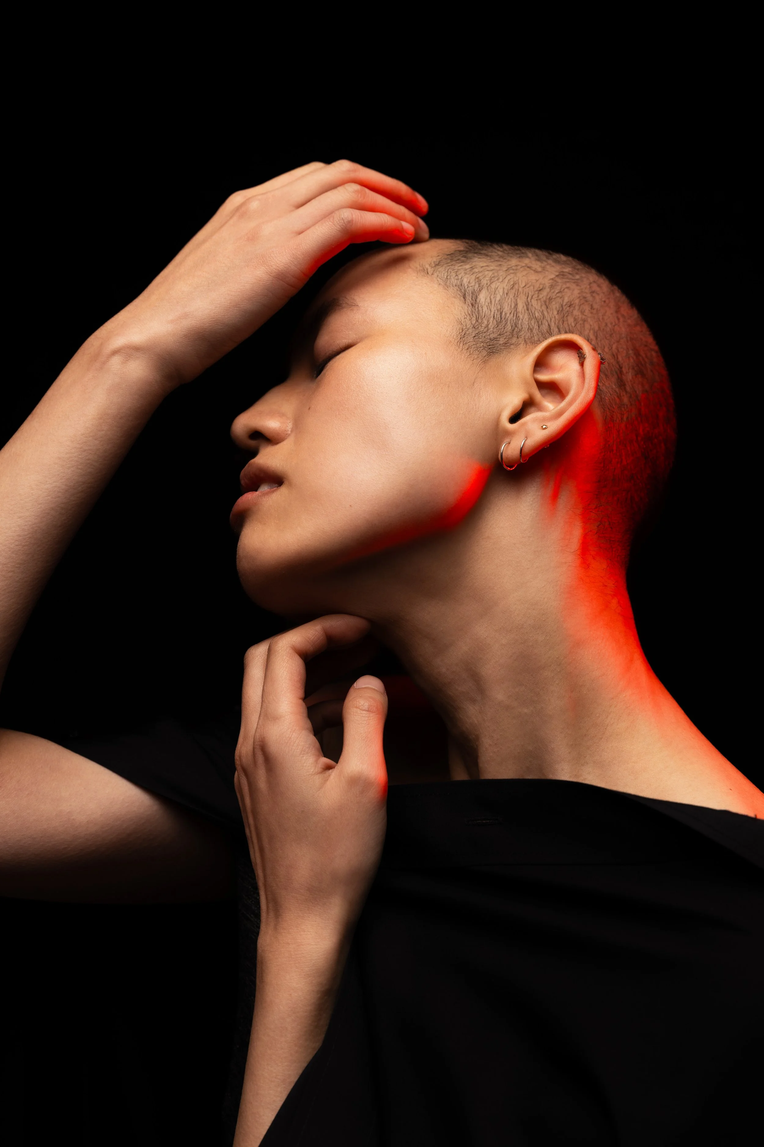 A person with a shaved hairstyle and earrings, posing against a black background with red lighting highlighting the side of the face and neck, eyes closed, hand on forehead and chin.